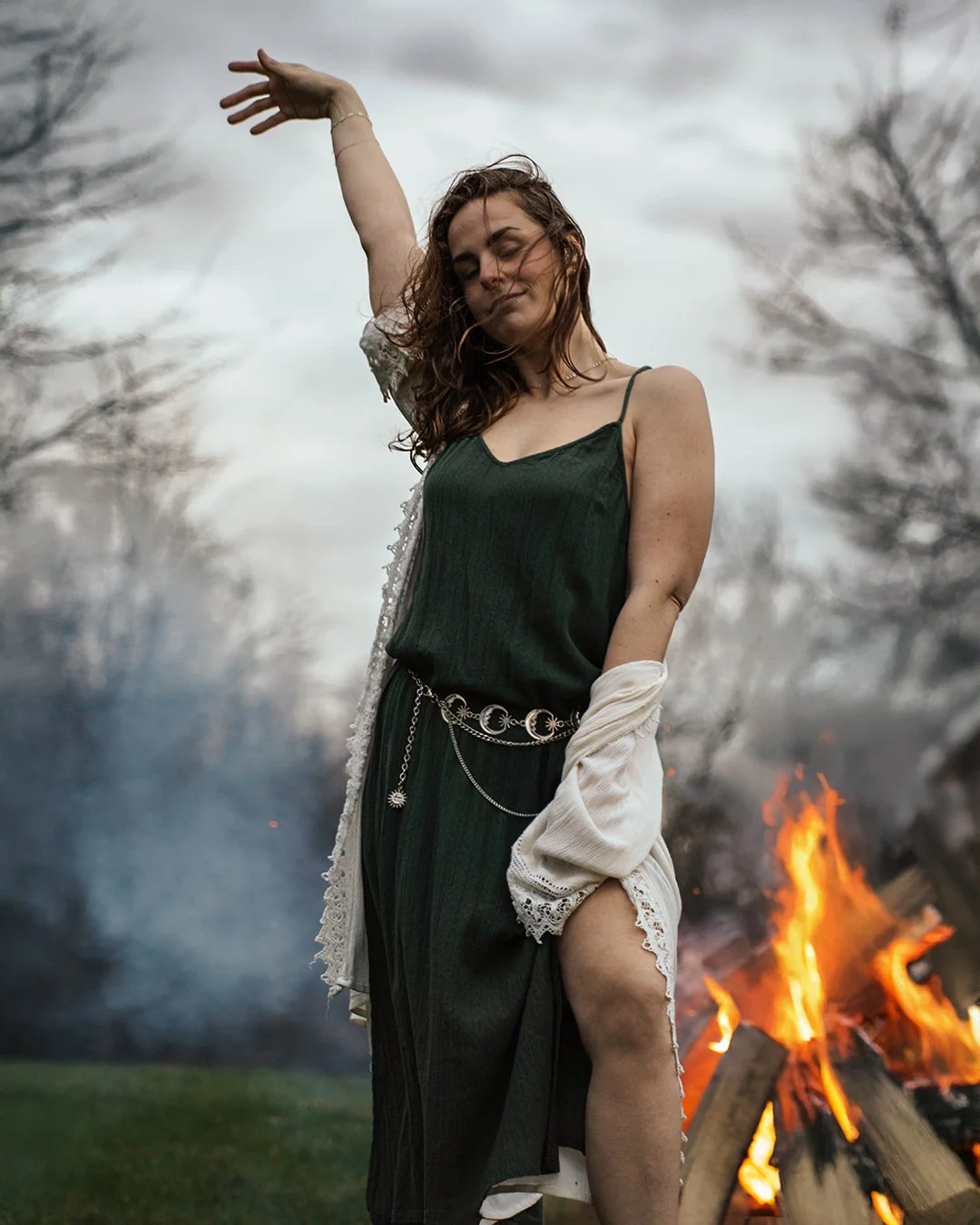A woman with curly hair dancing in front of a bonfire outdoors on a cloudy day.