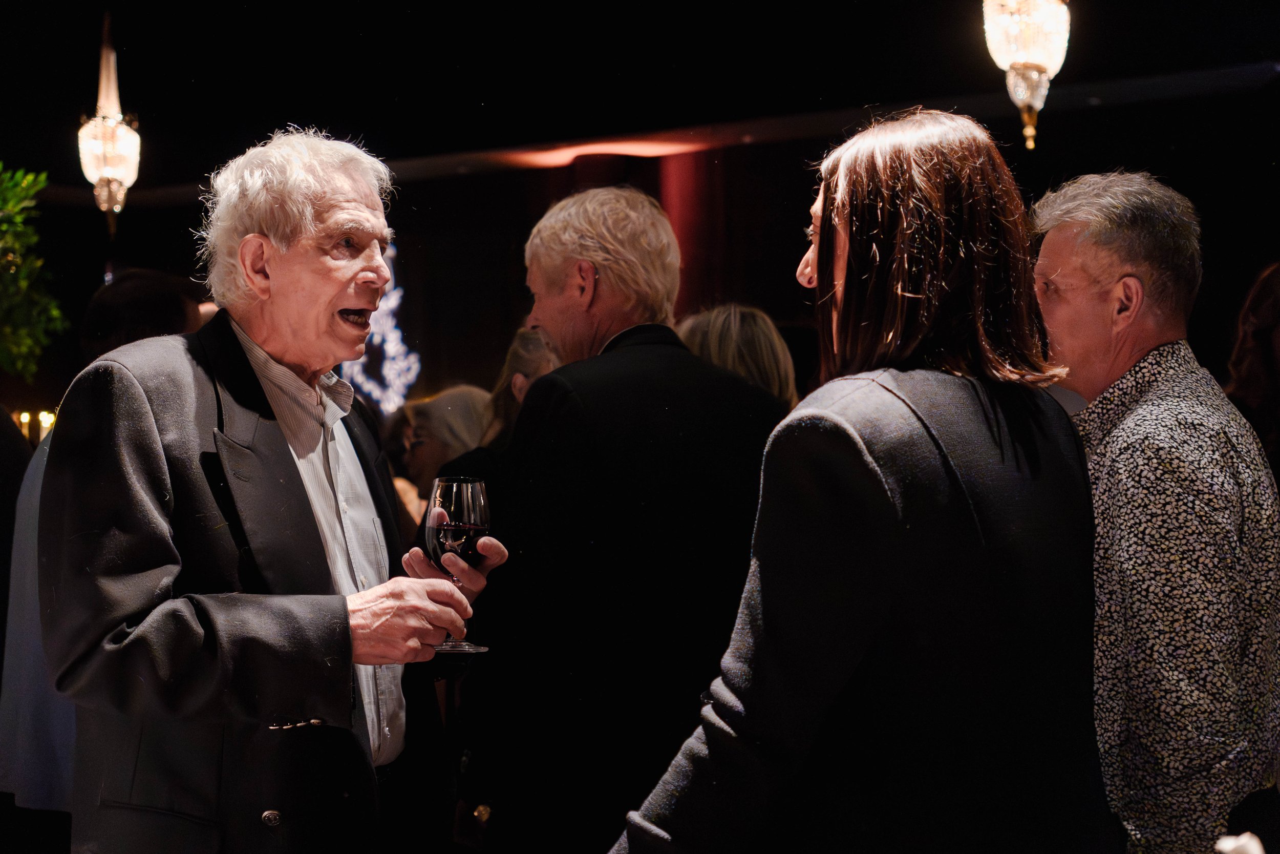 Group of people at a social gathering, engaged in conversation, with one older man holding a glass of red wine.