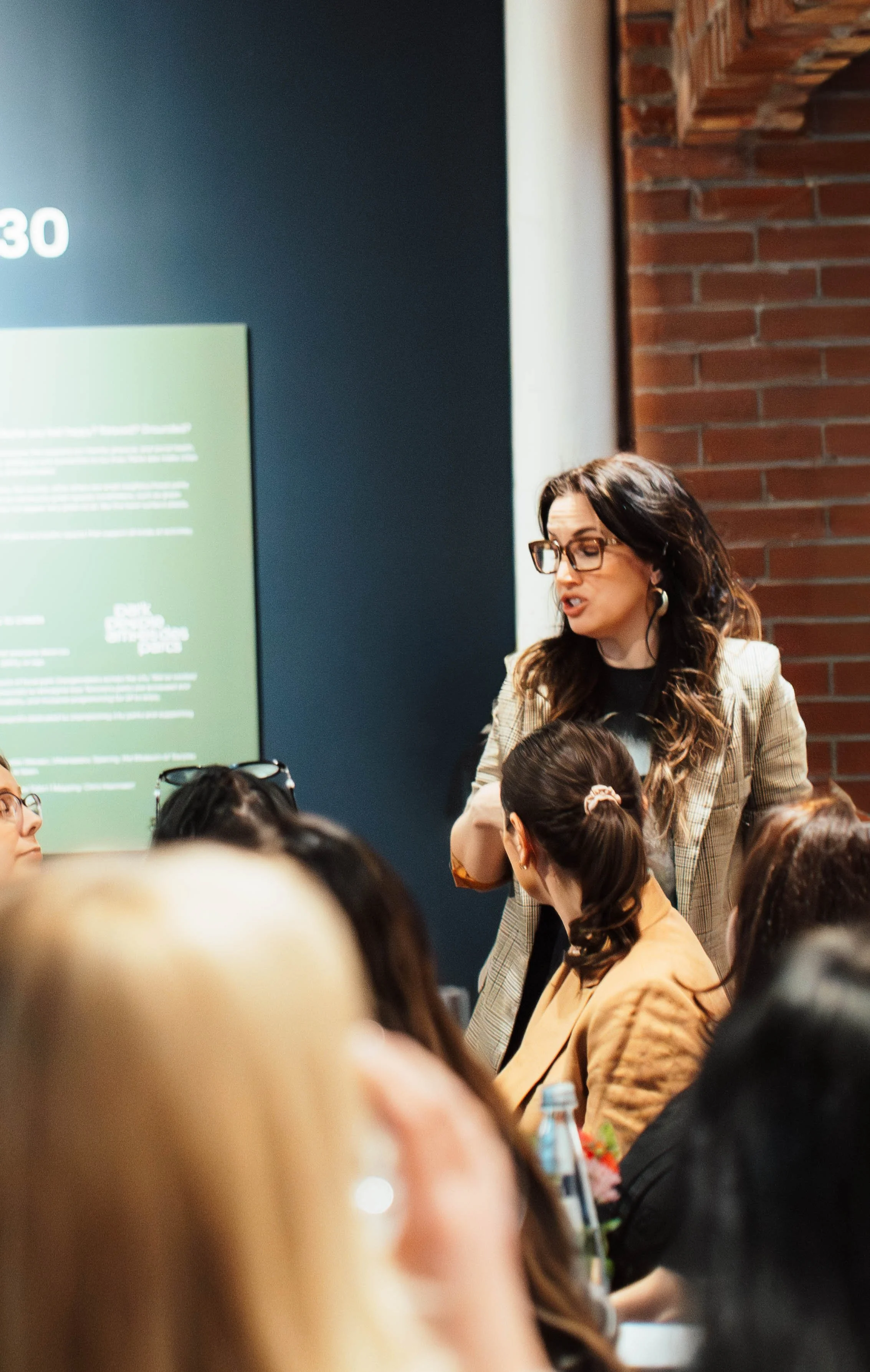 A woman with glasses and long hair speaking to a group of people, possibly at a conference or seminar, with a green information board and a brick wall in the background.
