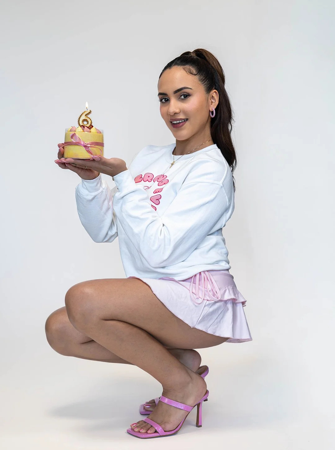 A woman with long dark hair tied in a ponytail, wearing pink high-heeled sandals, a white sweatshirt with pink lettering, and a pink skirt or shorts, is kneeling and holding a small birthday cake with a lit candle shaped as the number 6, celebrating 