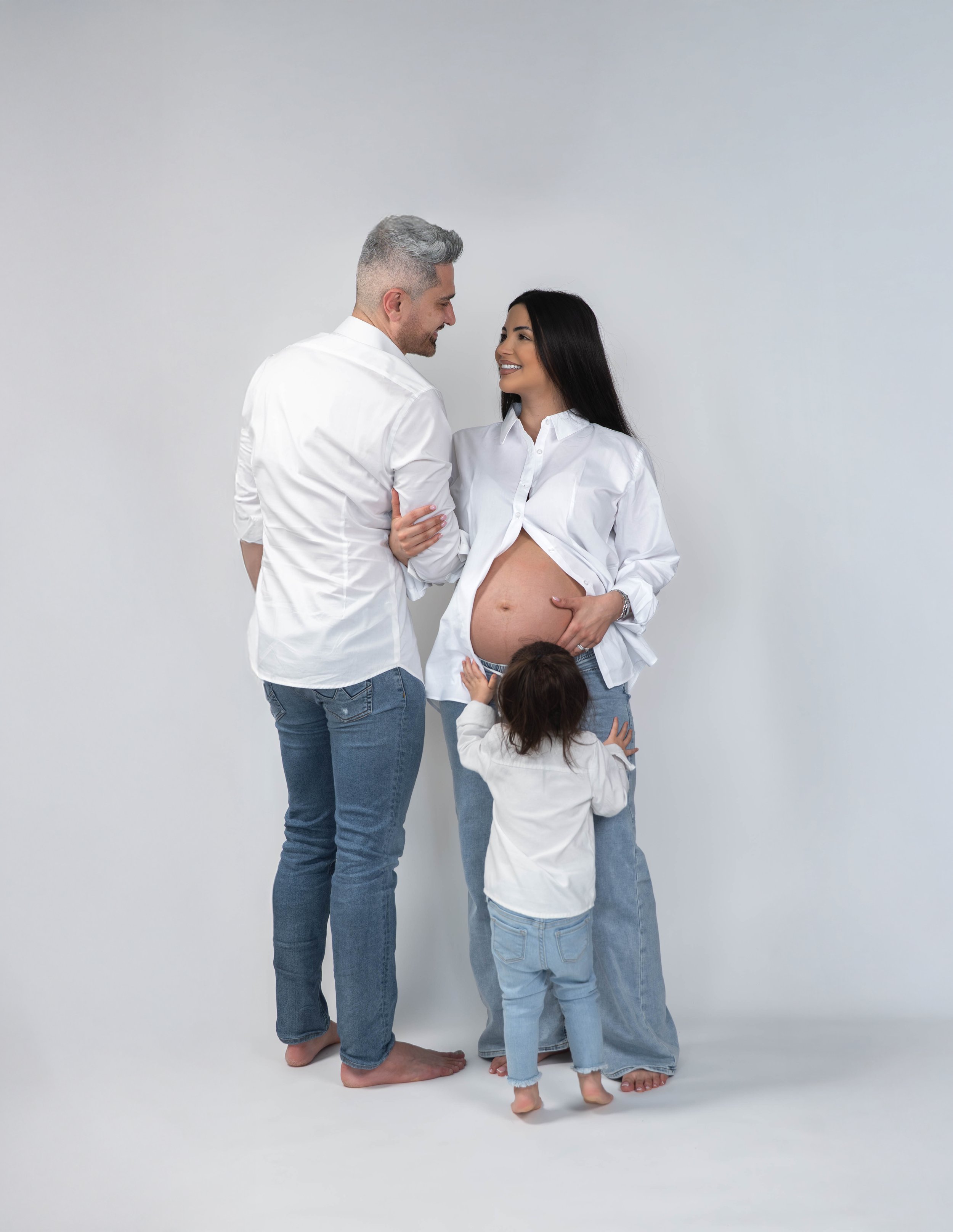 A couple, a man and a woman, are standing barefoot on a plain white background. The woman is pregnant and is wearing a white shirt, with her hand resting on her belly, and smiling at the man. The man is wearing a white shirt and blue jeans, and is sm