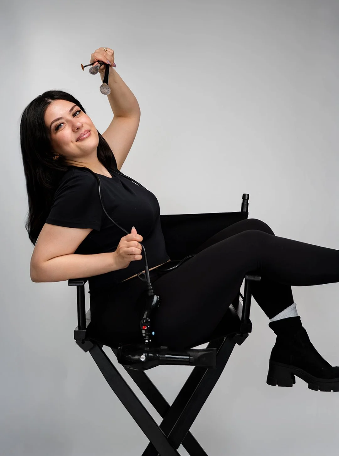 A young woman with black hair, wearing a black t-shirt, black pants, and black boots, sitting on a director's chair. She is smiling and holding a boom microphone above her head, with a wireless microphone pack attached to her pants.