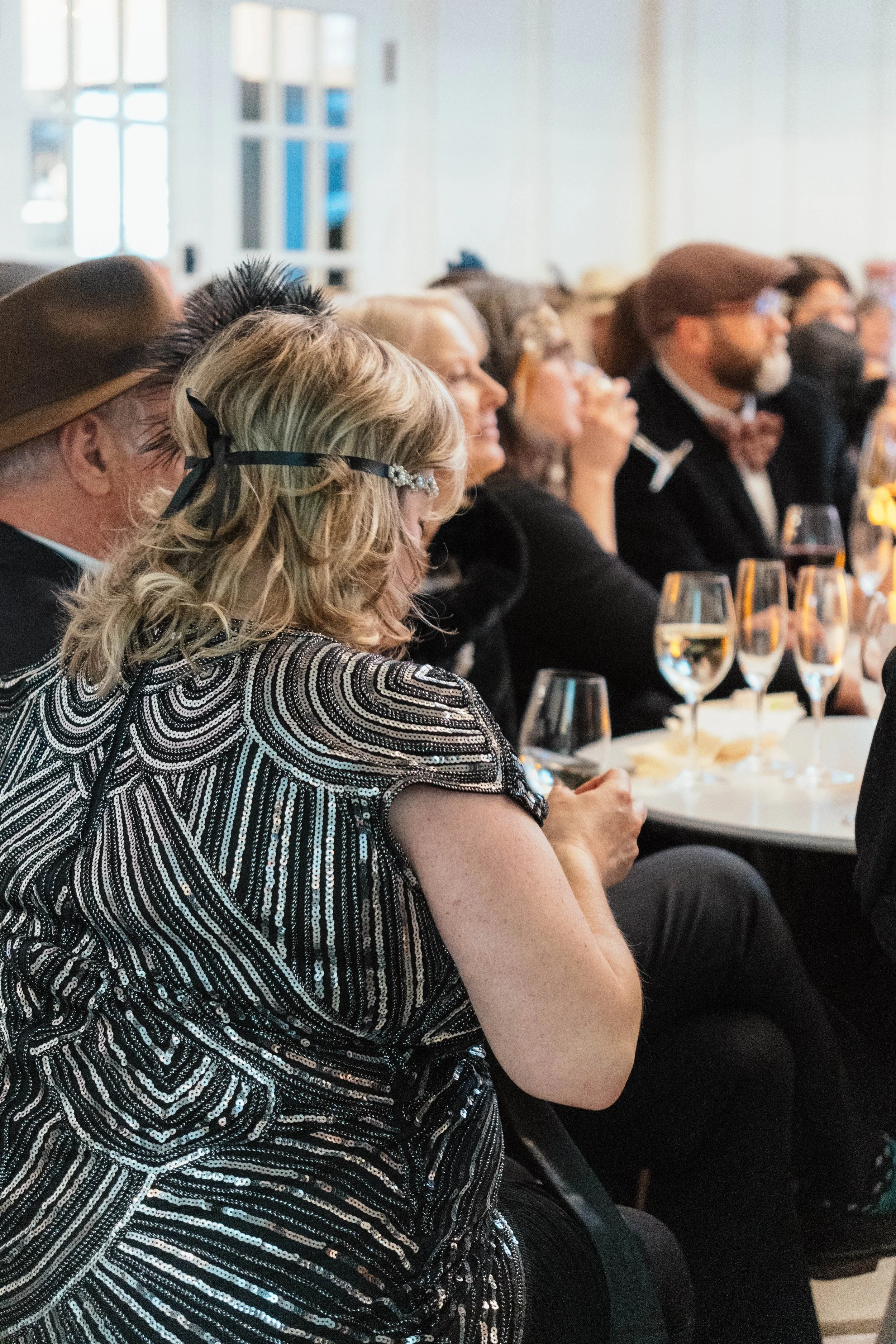 People seated at a formal event, some holding glasses of wine, paying attention to a speaker or presentation in a well-lit room.