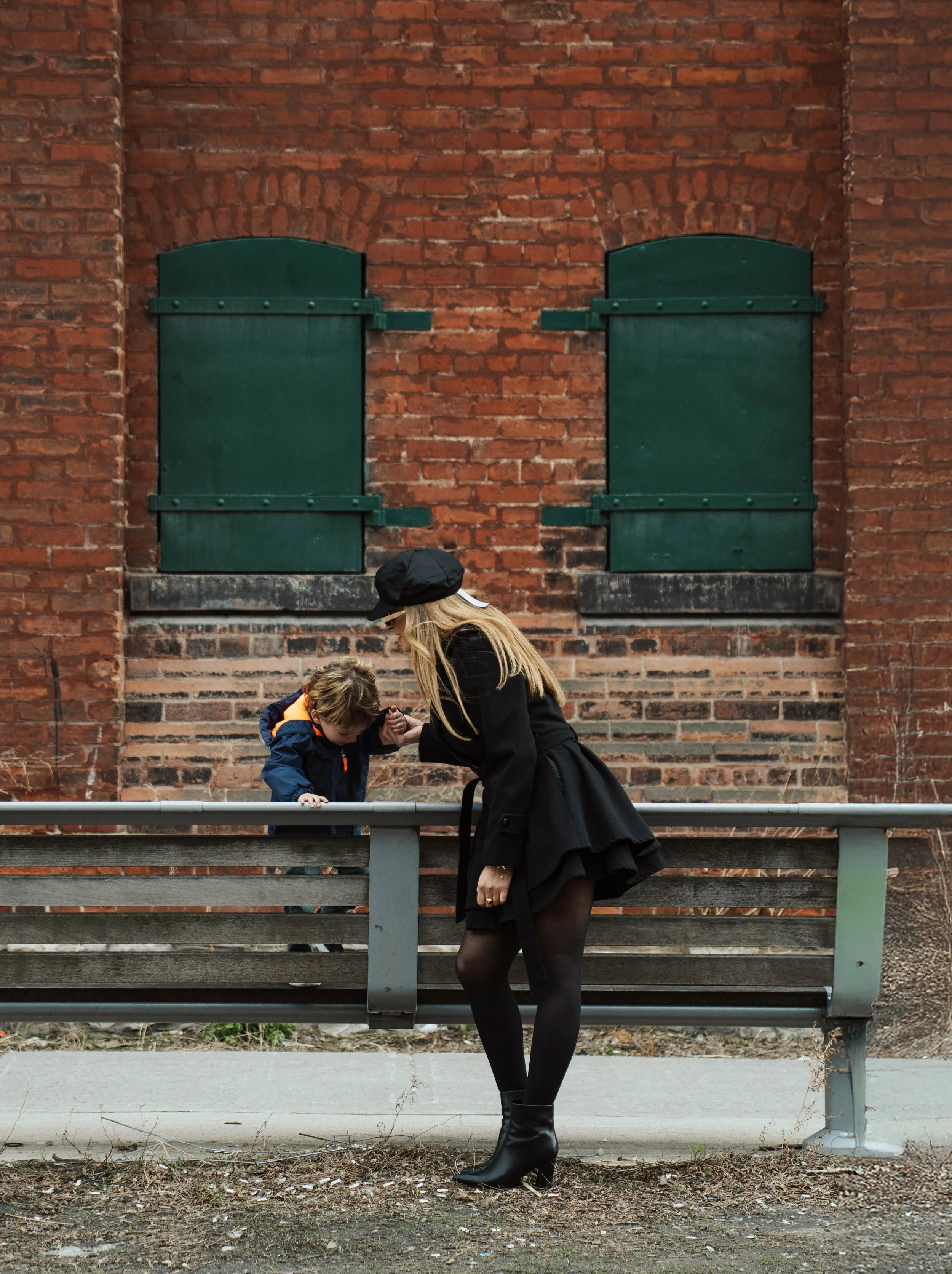 A woman dressed in black with high-heeled boots leaning down beside a young boy with light brown hair, who is looking at something on the ground, both standing next to a metal fence in front of a red brick wall with closed green shutters.