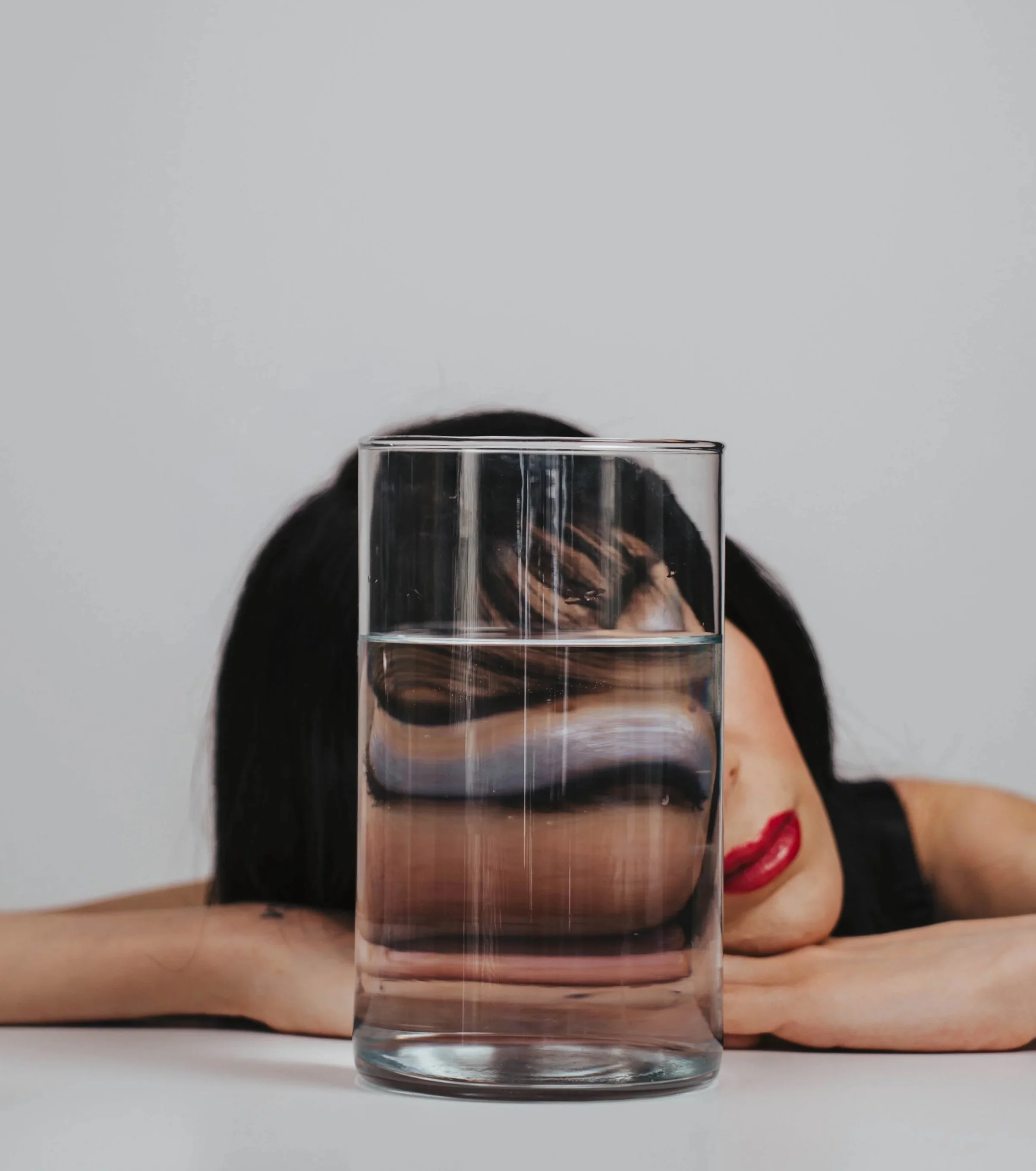 Woman creating surreal self portrait photography using glass and water refraction distortion in a minimalist studio setup with conceptual fine art portrait styling.