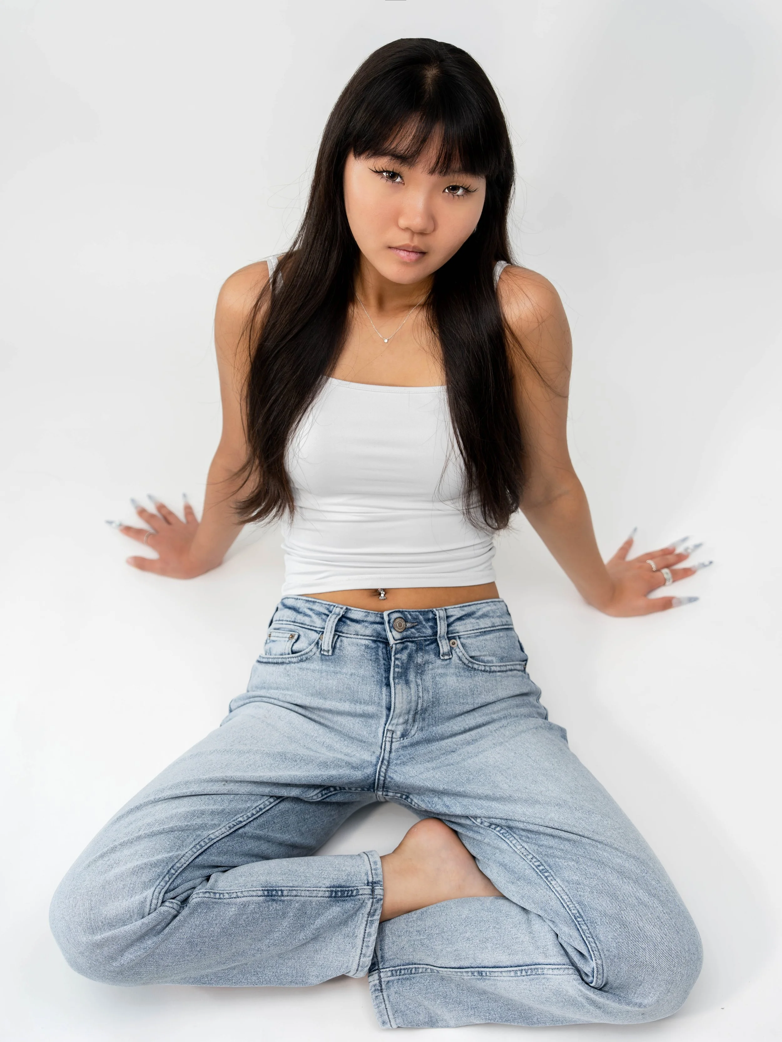 A young woman with long black hair and bangs is sitting on the floor in a white tank top and jeans, against a plain white background.