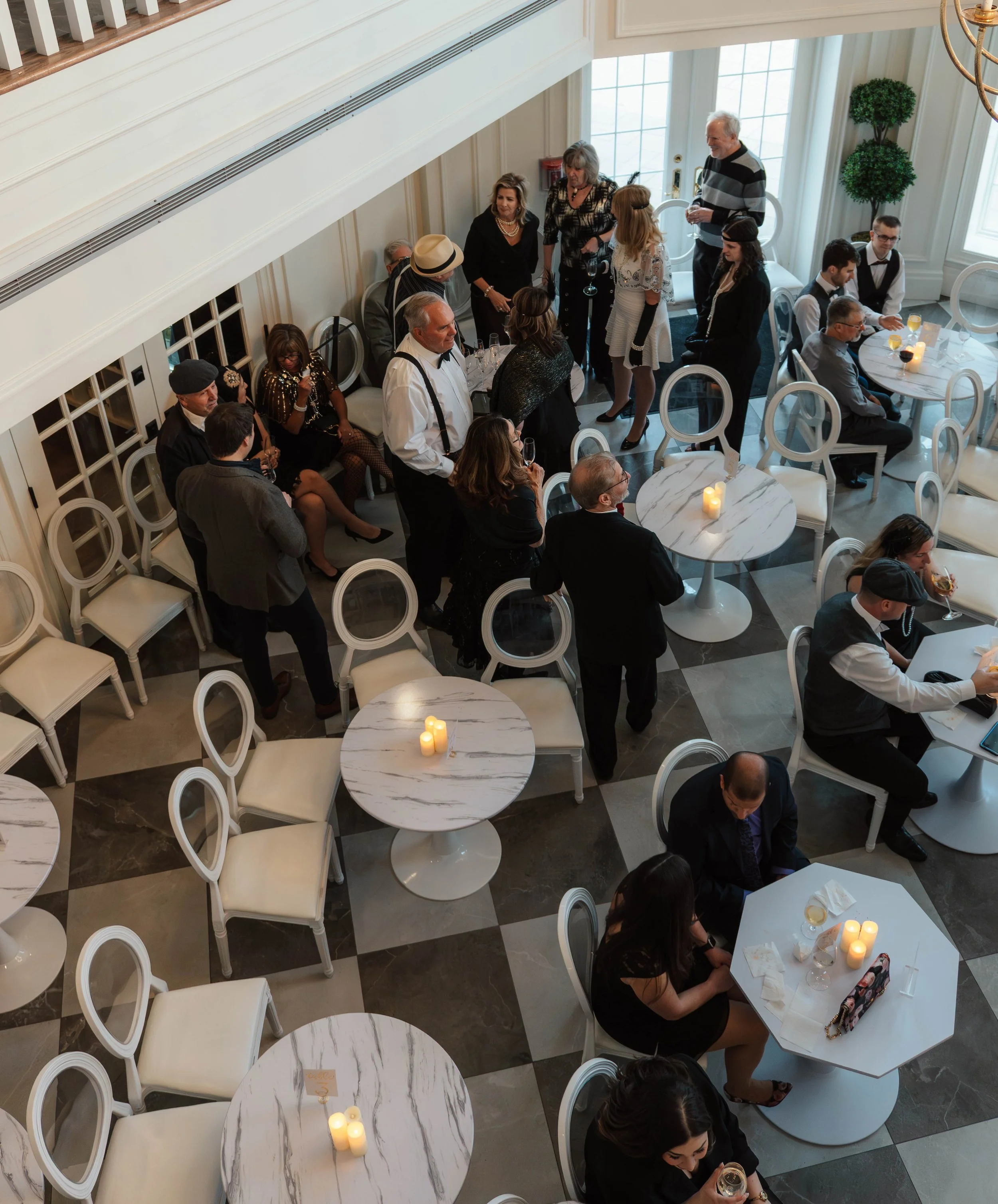 People gathered at a social event in a well-lit room with white walls, checkered black and white floor, white chairs, and candles on marble tables, some standing and some sitting, engaged in conversation or using devices.