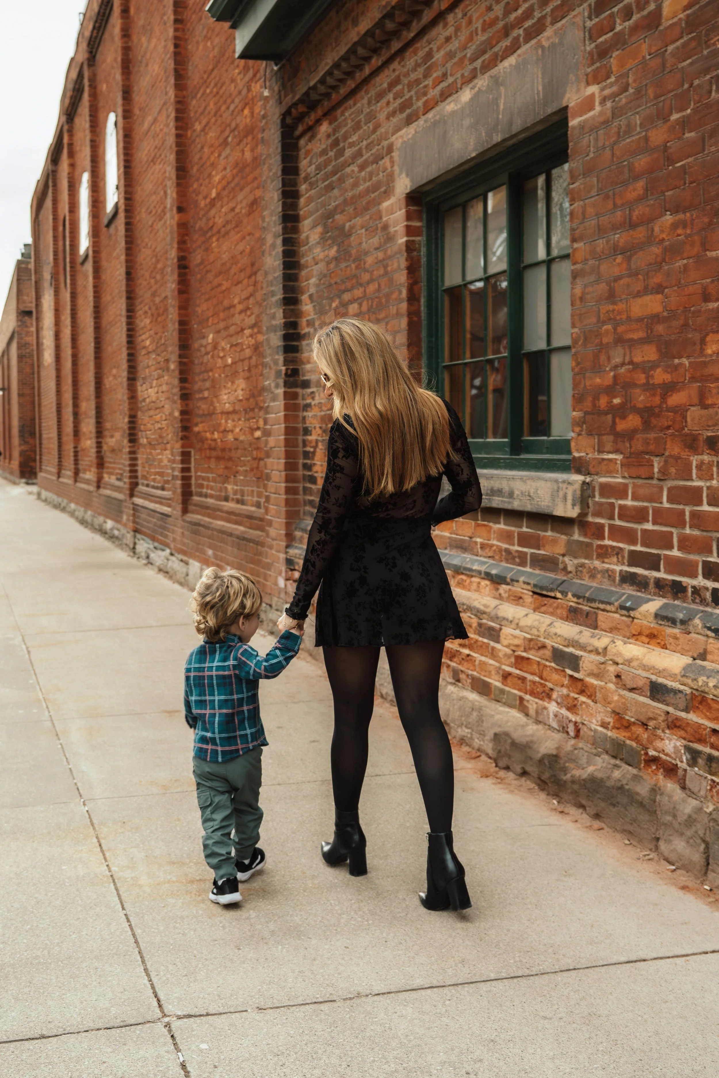 A woman with long blonde hair, wearing a black lace dress and black high-heeled boots, holding hands with a young boy in a blue plaid shirt and gray pants, walking along a sidewalk next to a red brick building with green window frames.