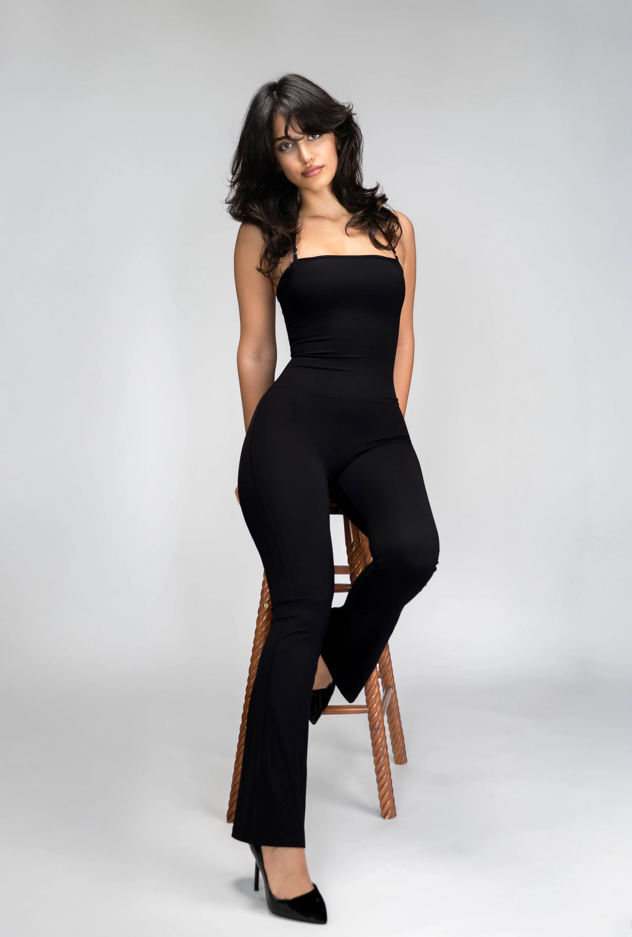 Woman seated on a wooden stool wearing an all-black outfit with black high heels, posing against a plain light gray background.