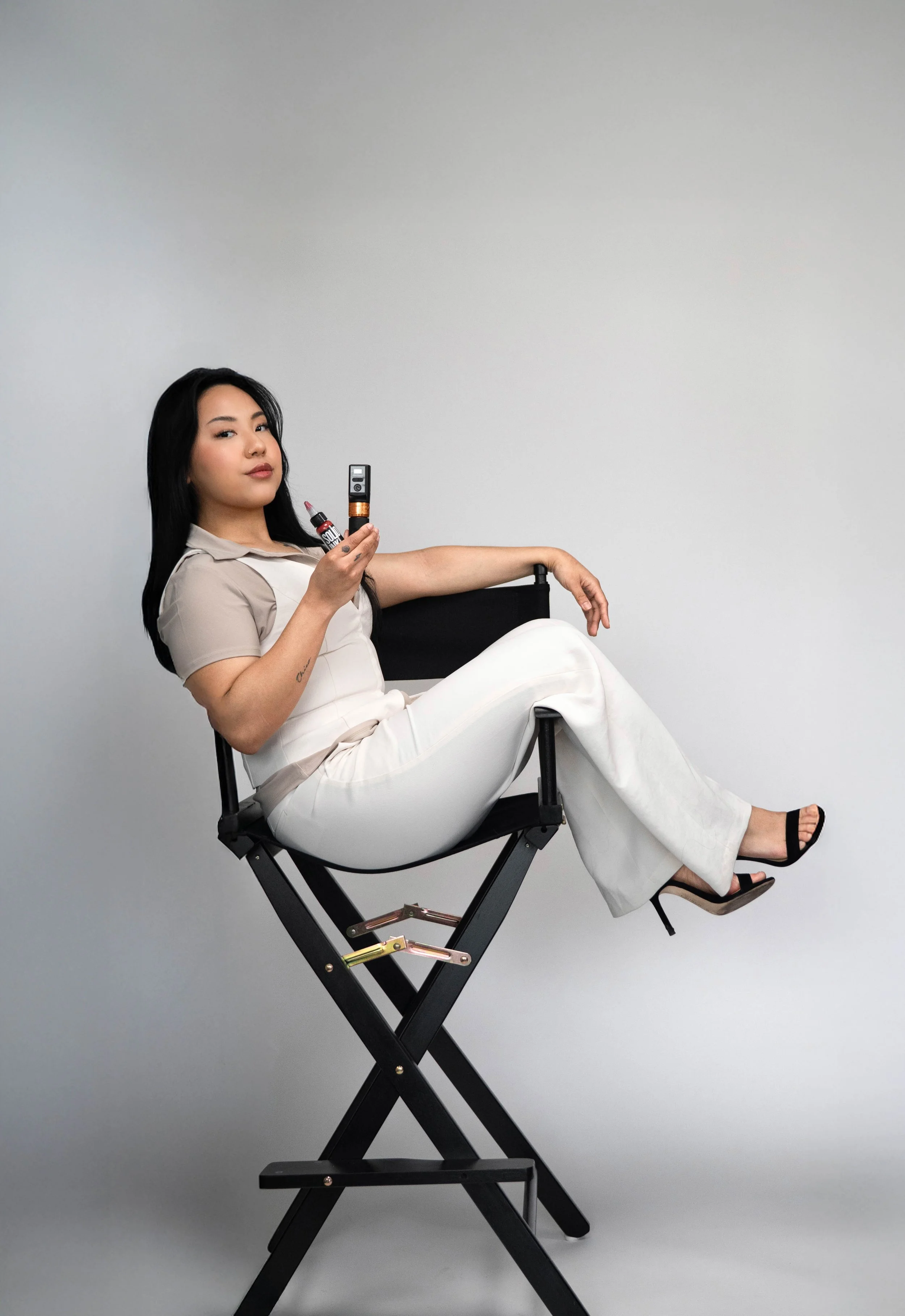 A woman with black hair, wearing a beige shirt and white pants, sits on a black director's chair against a plain gray background. She is holding a makeup brush in one hand and a compact mirror in the other, with her legs crossed and smiling softly.