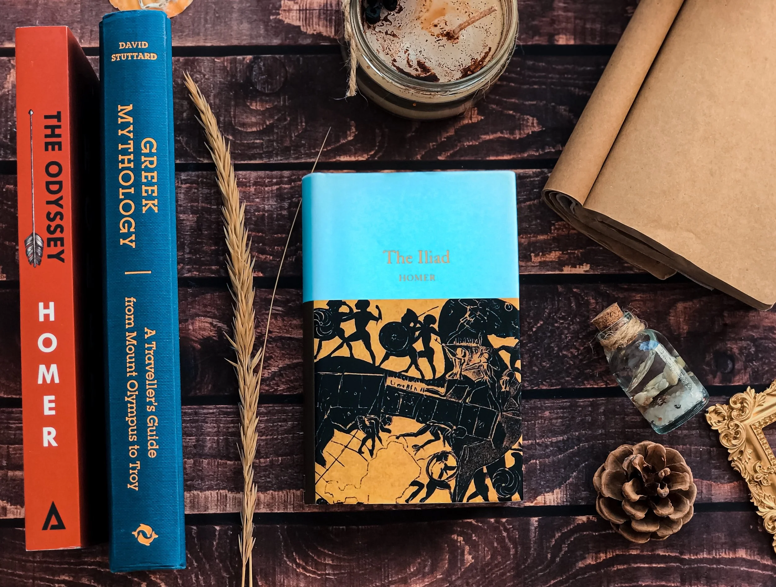 Arrangement of books, a polar bear figurine, a small glass jar with shells, a pine cone, a cardboard tube, a decorative frame, a glass jar with a candle, and a blue book titled 'The Iliad' on a dark wooden surface.