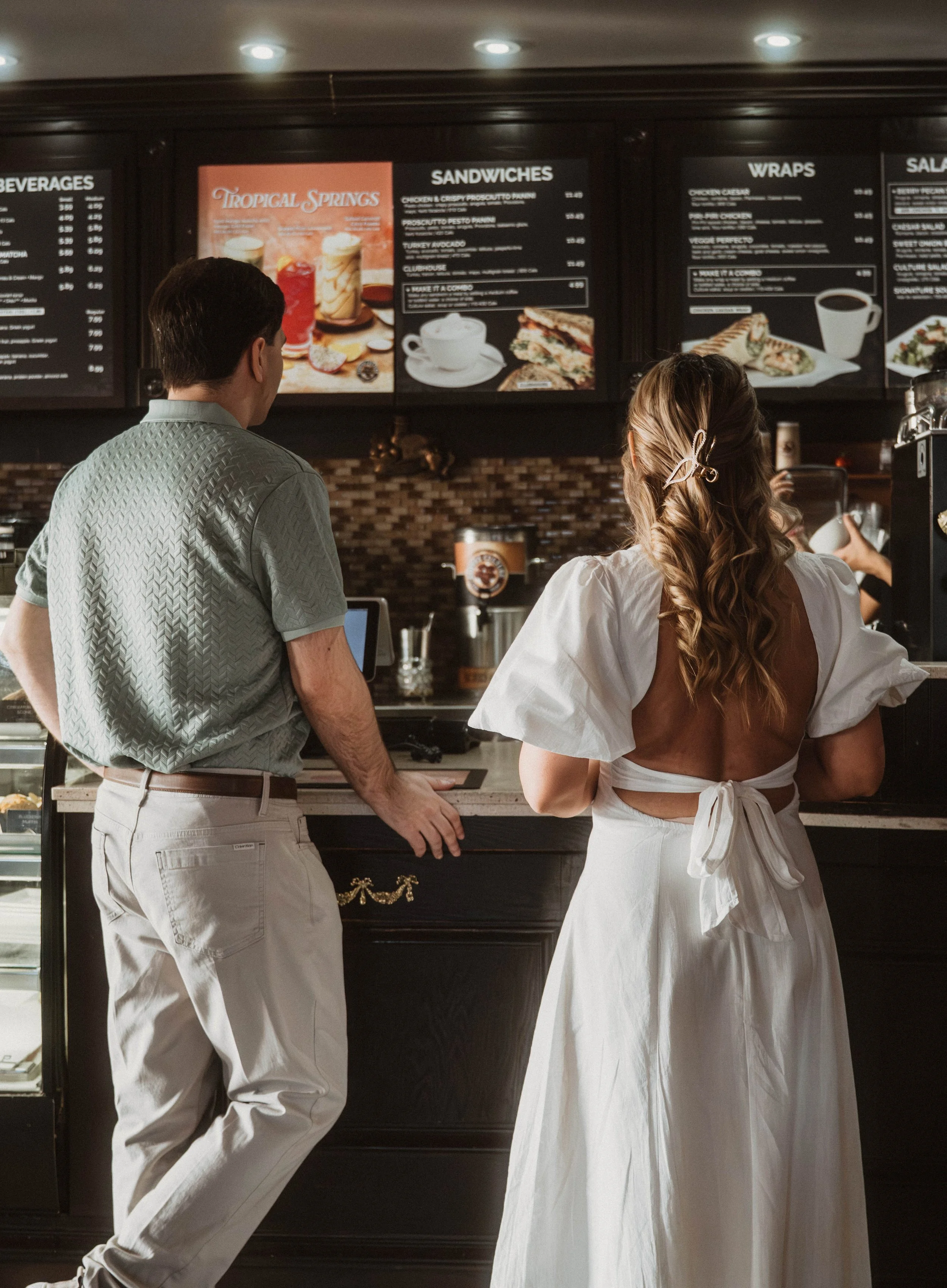 A man and a woman in a coffee shop, facing the menu board. The man is wearing white pants and a patterned shirt, and the woman is wearing a white dress with a large bow on the back, her hair styled with a hair clip. The menu board displays various fo