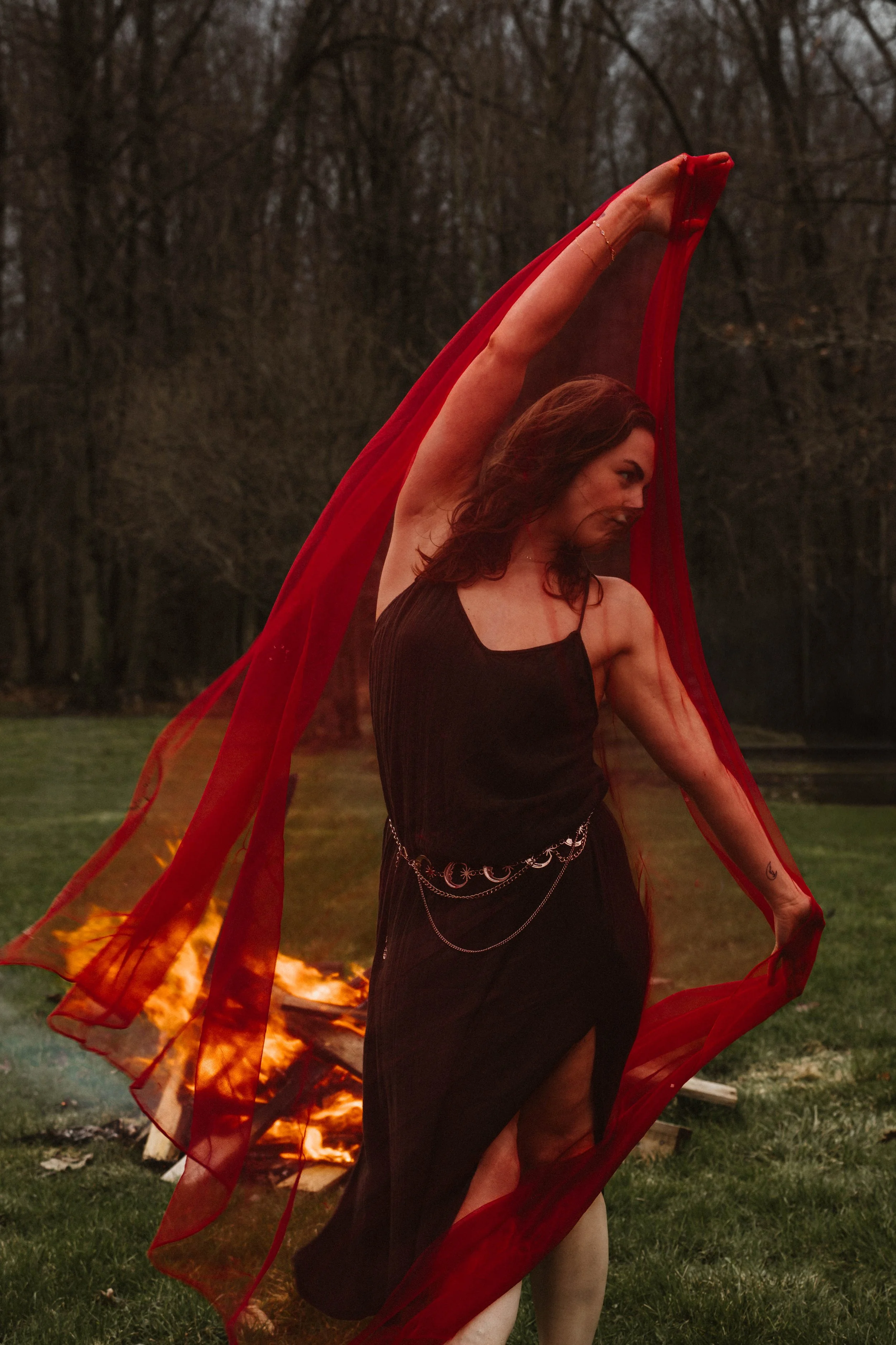 A woman in a black dress with chain accessories poses with red sheer fabric, in front of a fire outdoors during dusk, with a background of trees.