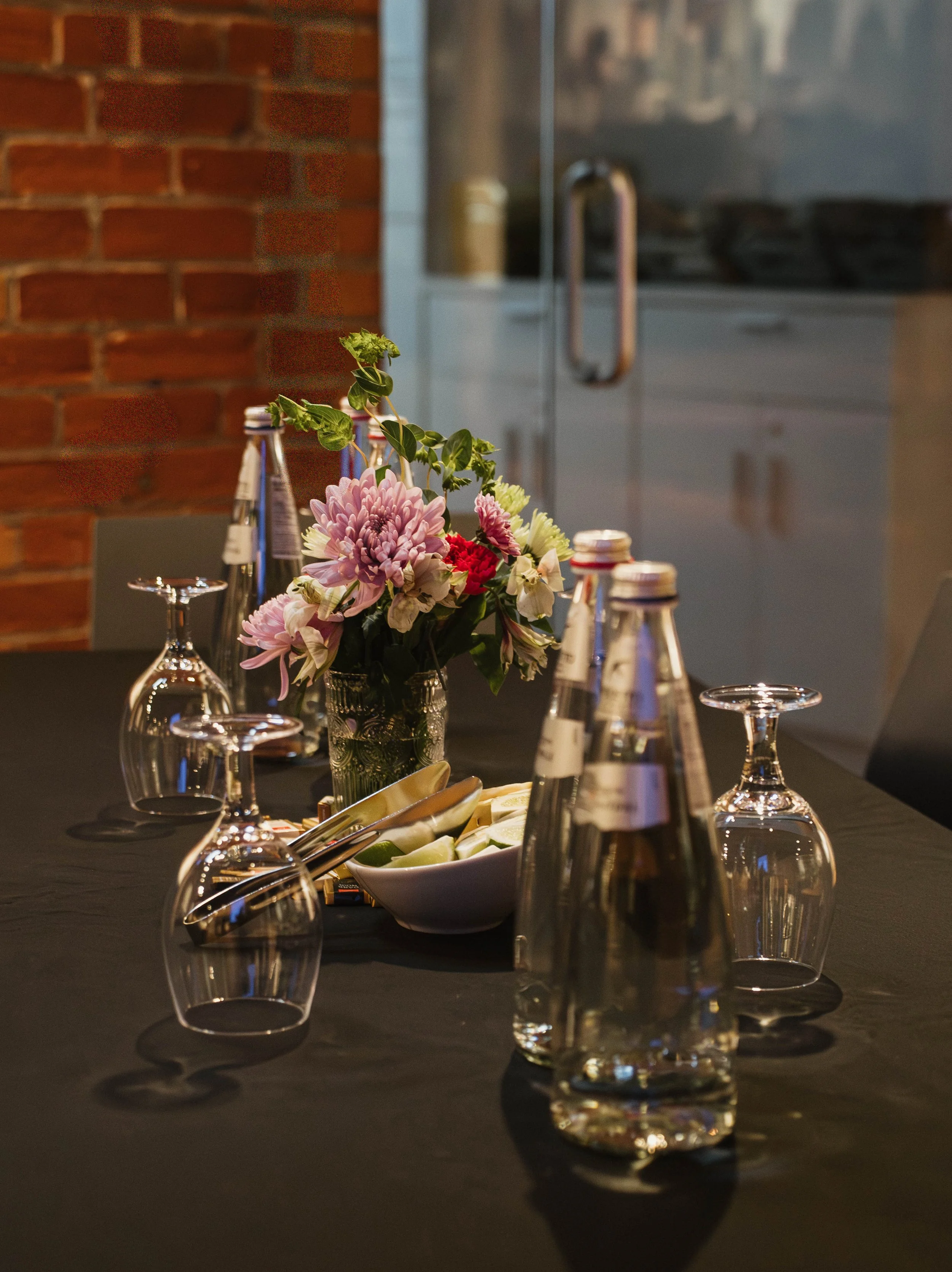 A dining table set with upside-down wine glasses, bottles of water, a bowl of lime wedges, and a bouquet of flowers in a jar, against a backdrop of brick wall and kitchen appliances.