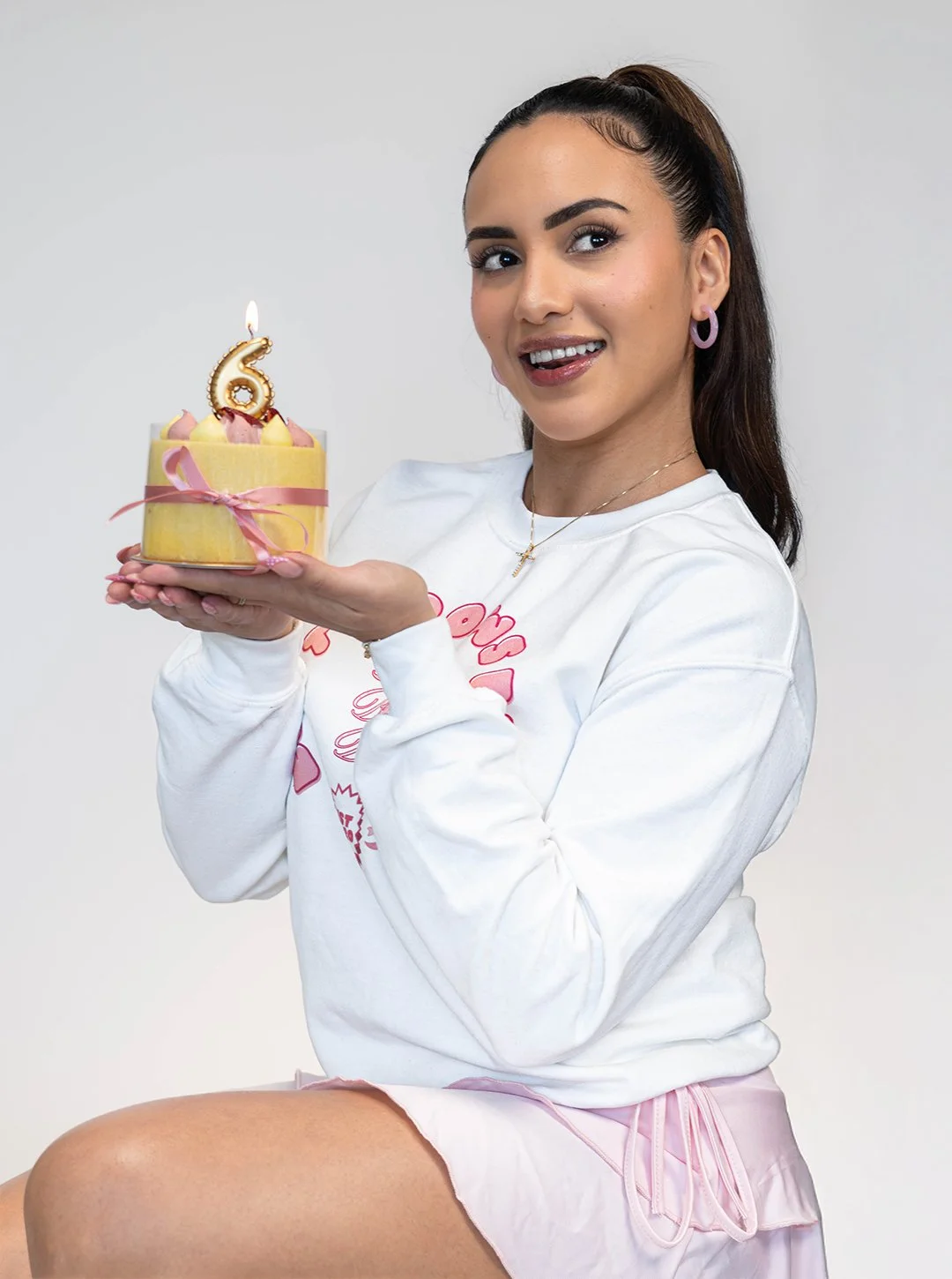 A young woman with dark hair tied back holding a small birthday cake with a candle shaped like the number 6. She is smiling and wearing a white sweatshirt with pink lettering and pink shorts.
