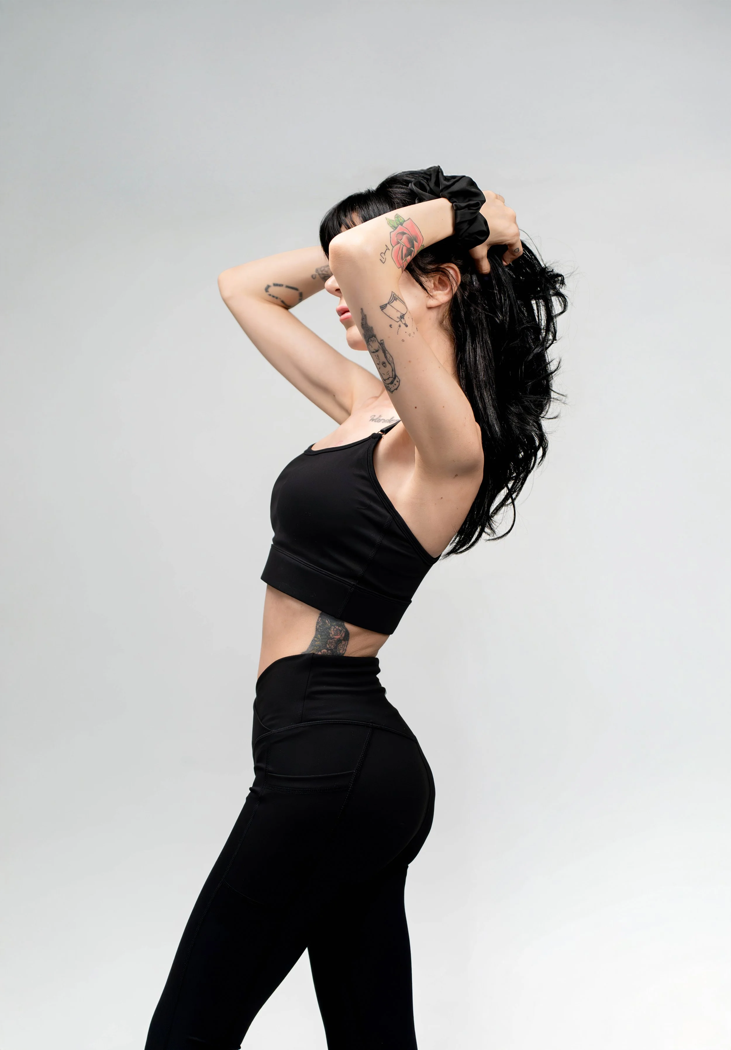 A woman with tattoos wearing a black sports bra and black leggings, stretching with her hands behind her head, against a plain light gray background.