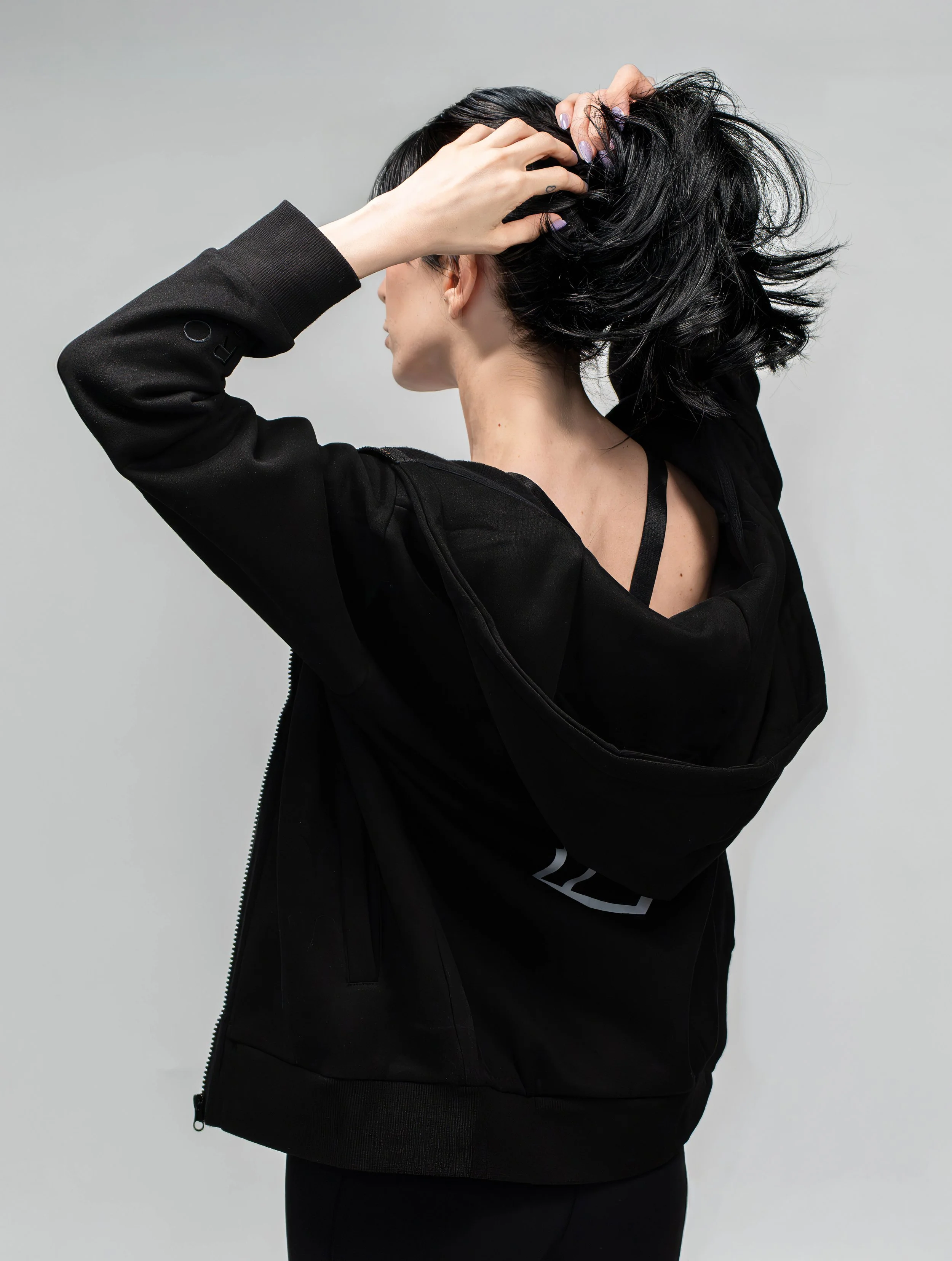 A woman with short black hair wearing a black hoodie, holding her hair back with one hand, turned slightly away from the camera.