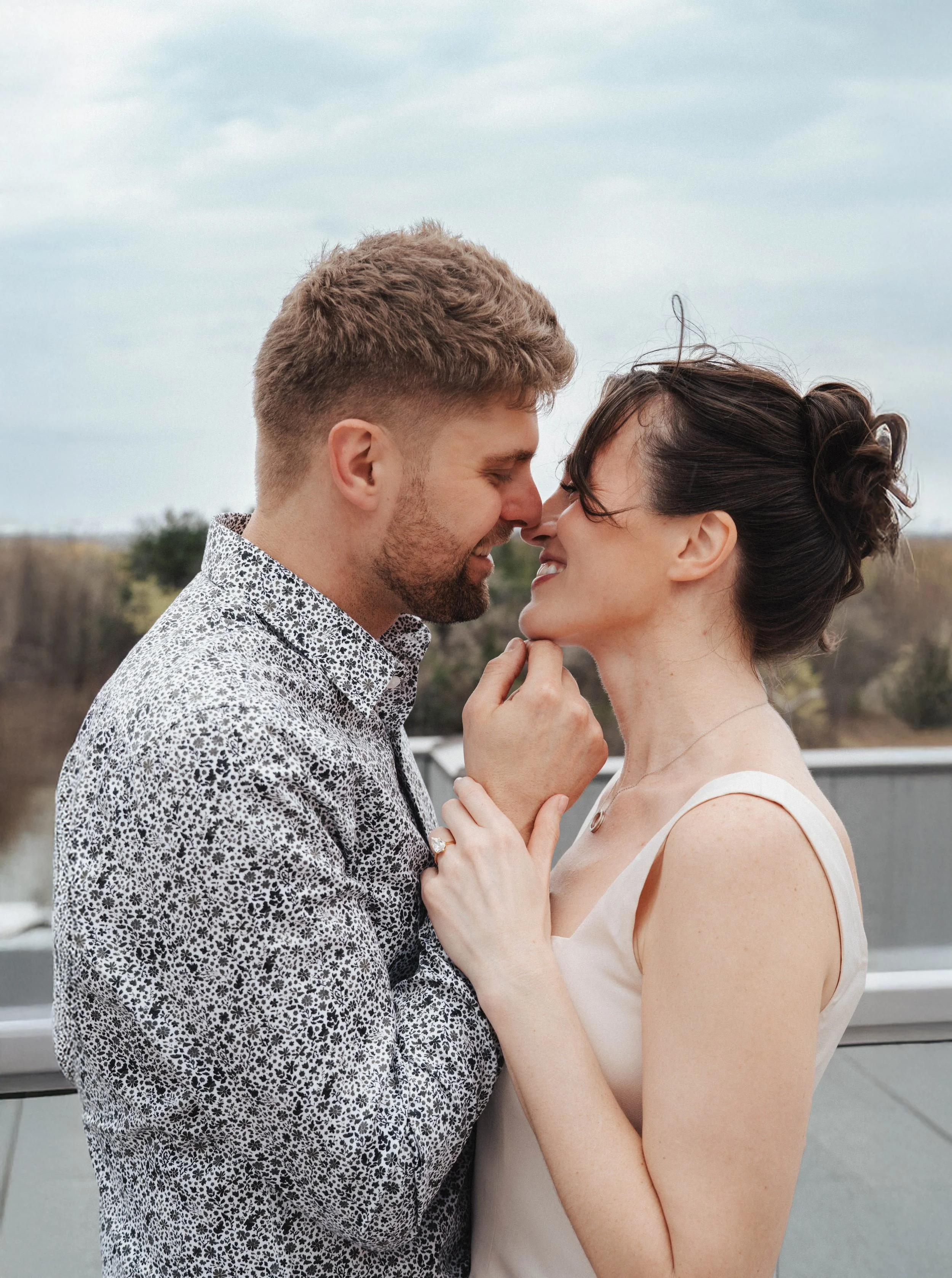 A couple faces closely together, smiling with eyes closed, standing outdoors on a cloudy day. The man has short, light brown hair and a patterned shirt. The woman has dark hair styled in an updo, and is wearing a sleeveless white dress with a ring on