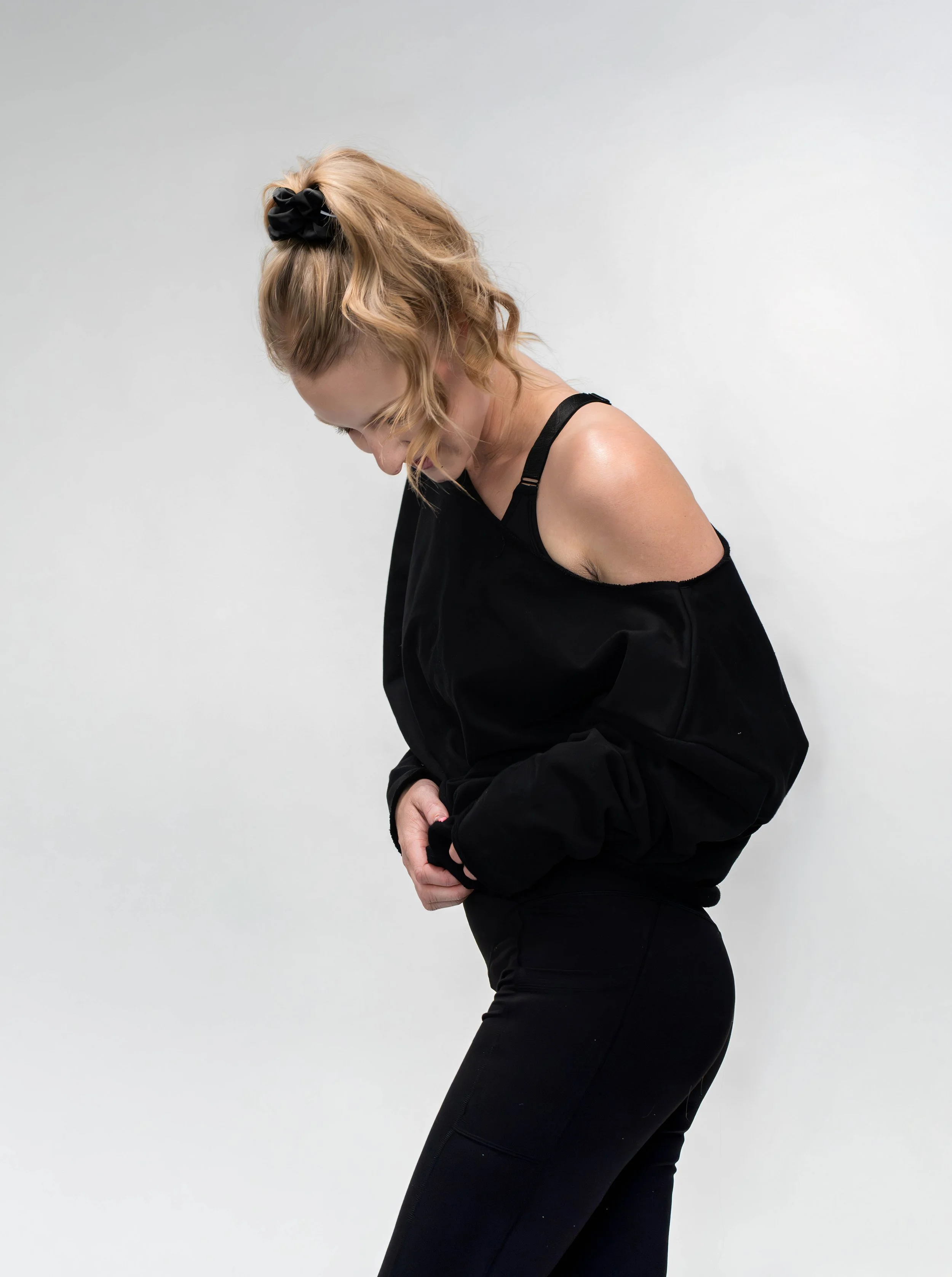 A woman with red hair tied back with a black scrunchie, wearing an off-shoulder black sweater and black pants, looking down against a plain white background.