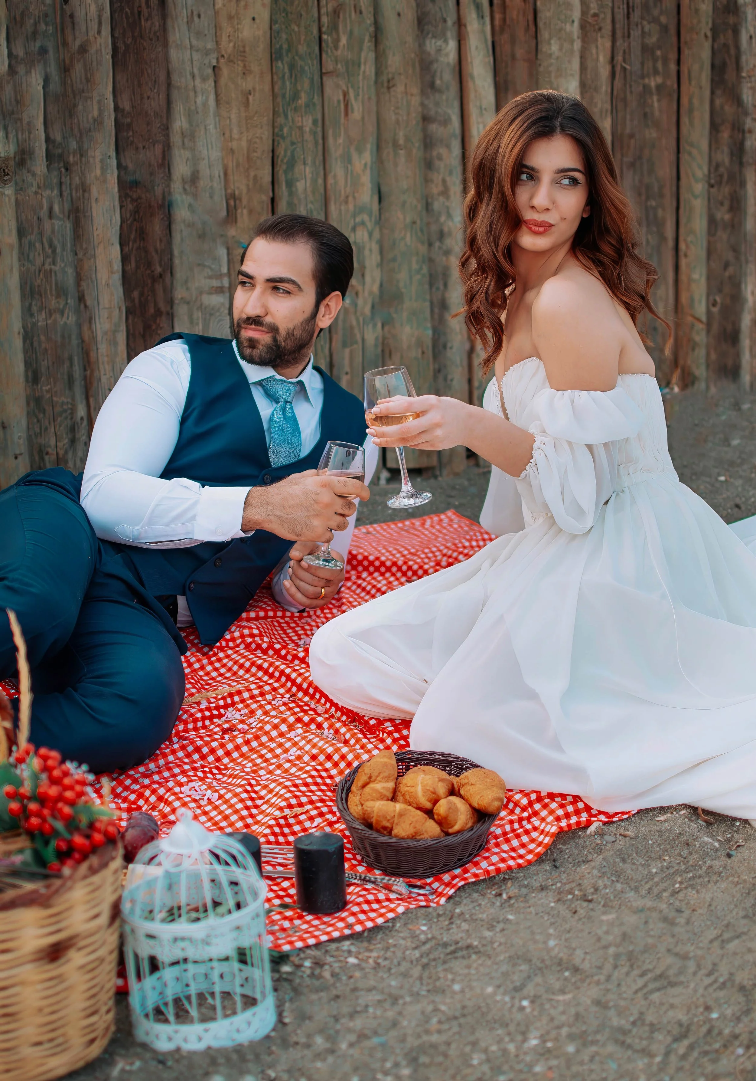 A man and woman having a picnic outdoors, sitting on a red gingham blanket with food and drinks, with a wooden fence in the background.