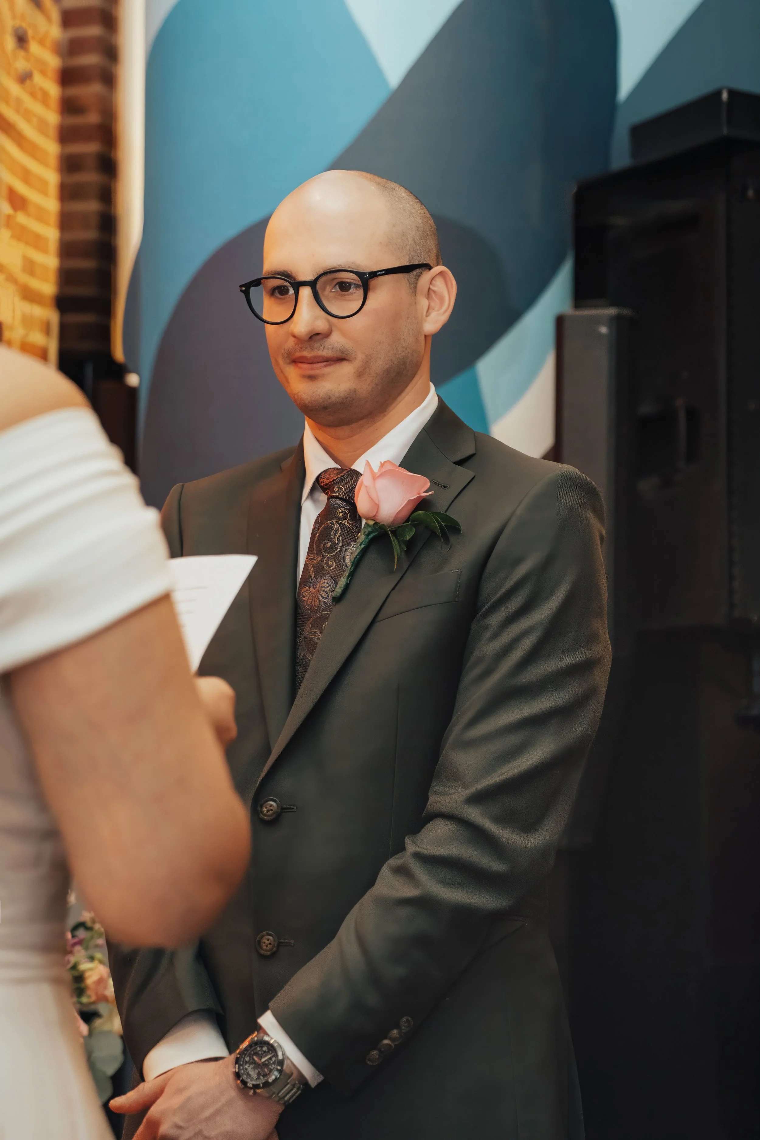 A man in a suit with a boutonnière, glasses, and a watch, standing at a ceremony or formal event.