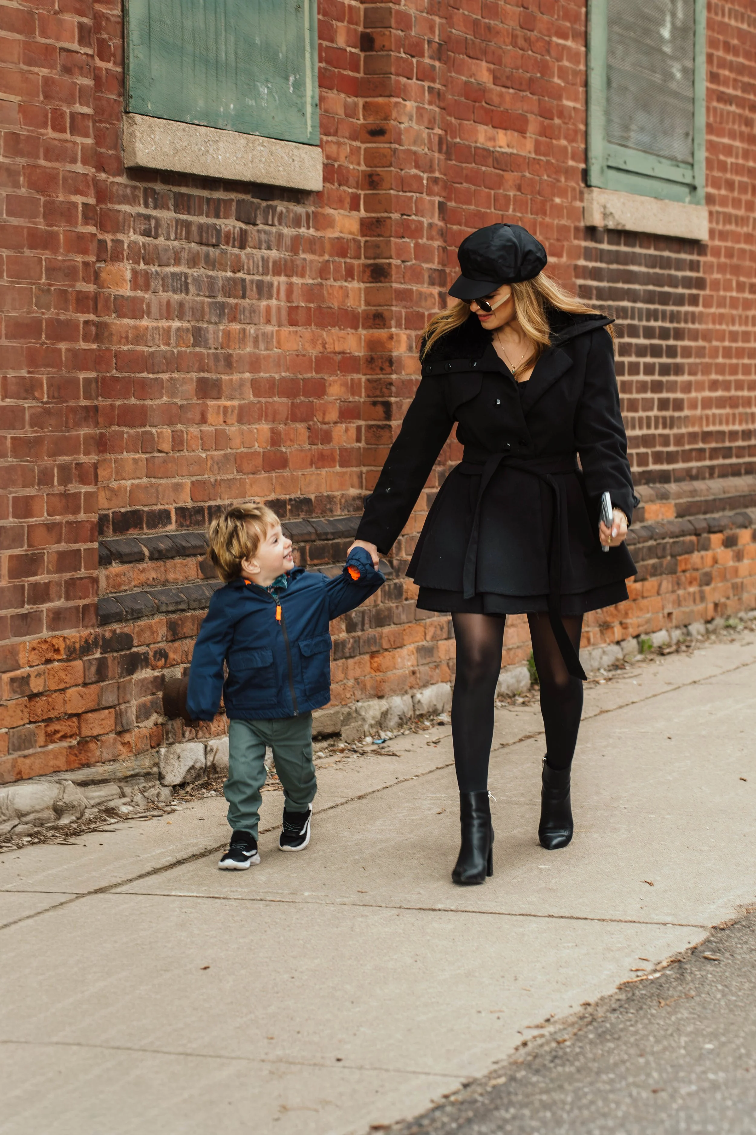 A woman dressed in black with tall boots and sunglasses holding hands with a young boy in a blue jacket and gray pants walking past a brick building on a sidewalk.