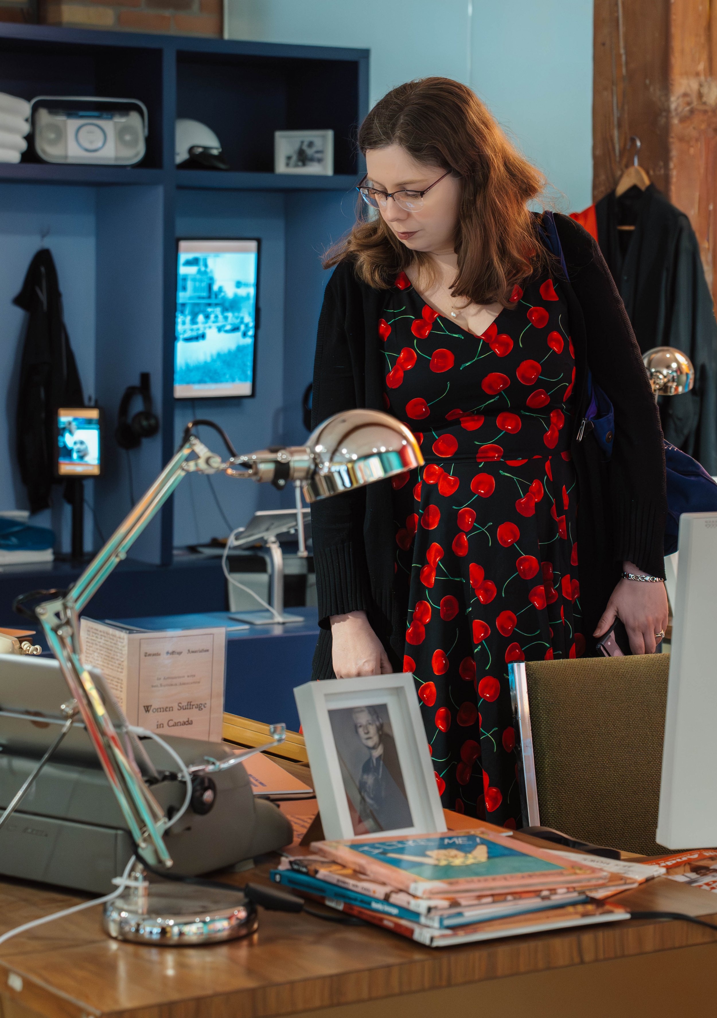 A woman wearing glasses and a black dress with red cherries stands at a desk in an office or museum setting. The desk has photographs, books, and a desk lamp. Shelves with various items, including a framed photo, a helmet, and a speaker, are in the b