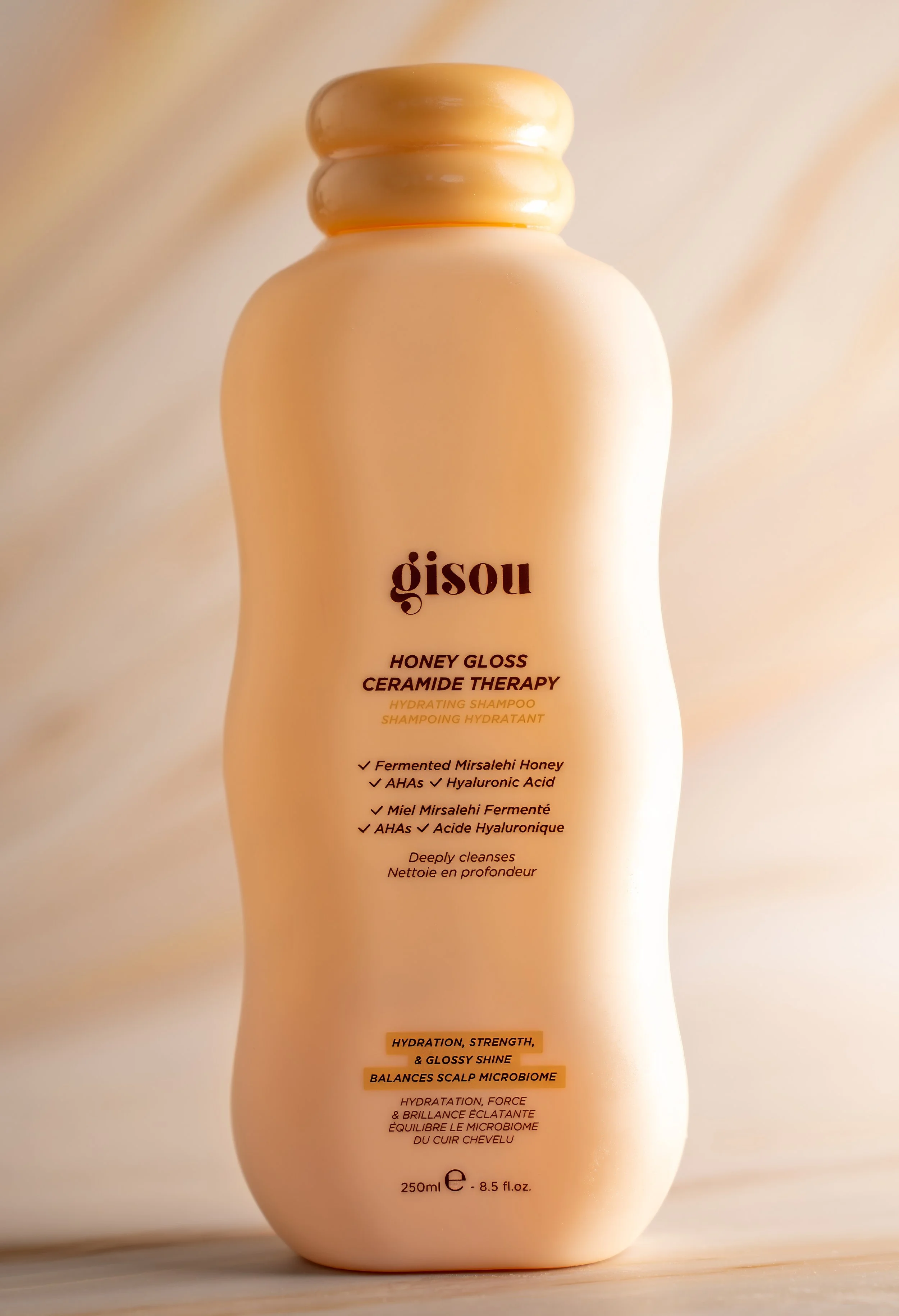 A beige bottle of Gisou Honey Gloss Ceramide Therapy hydrating shampoo with a rounded cap and minimalist design, placed against a soft, neutral background.