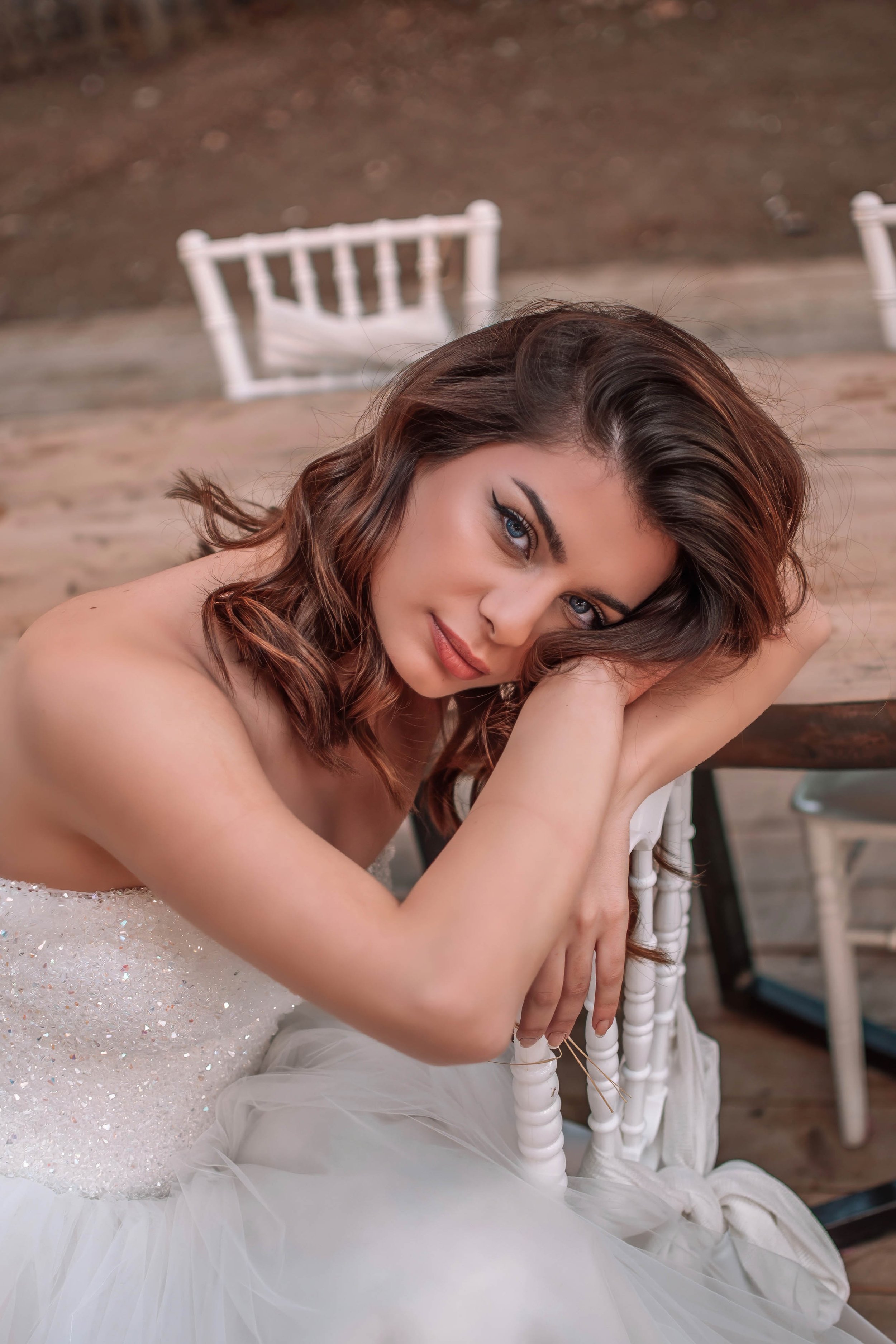 A woman with wavy brown hair and blue eyes sitting on a white chair, resting her head on her arms with a gentle smile. She is wearing a sparkly white dress and is in an outdoor setting with wooden flooring and a blurred background.