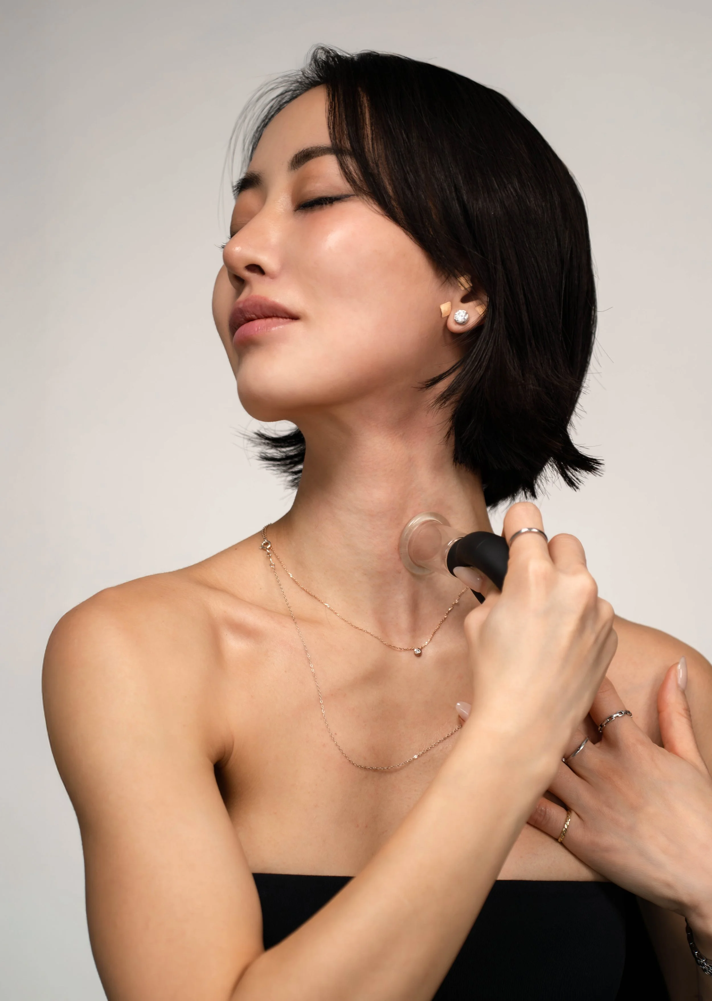A woman with short dark hair uses a facial cleansing device on her neck, wearing multiple delicate necklaces and earrings, with a serene expression and closed eyes.