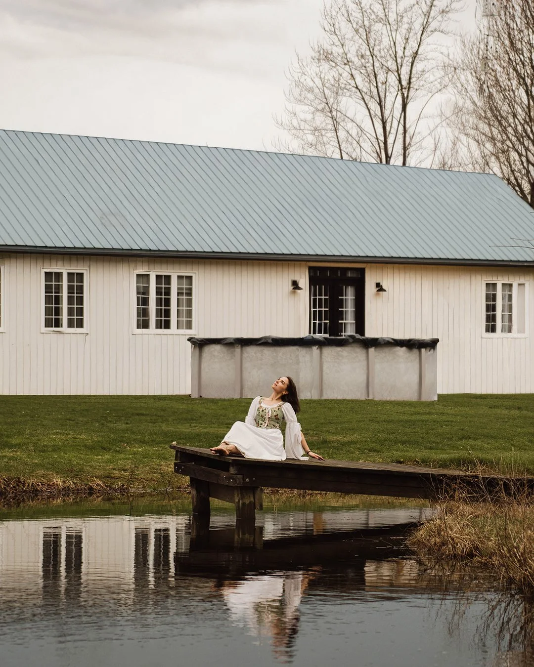 A woman in a white dress sits on a wooden dock by a calm pond, with a white barn with a light blue metal roof in the background and leafless trees.