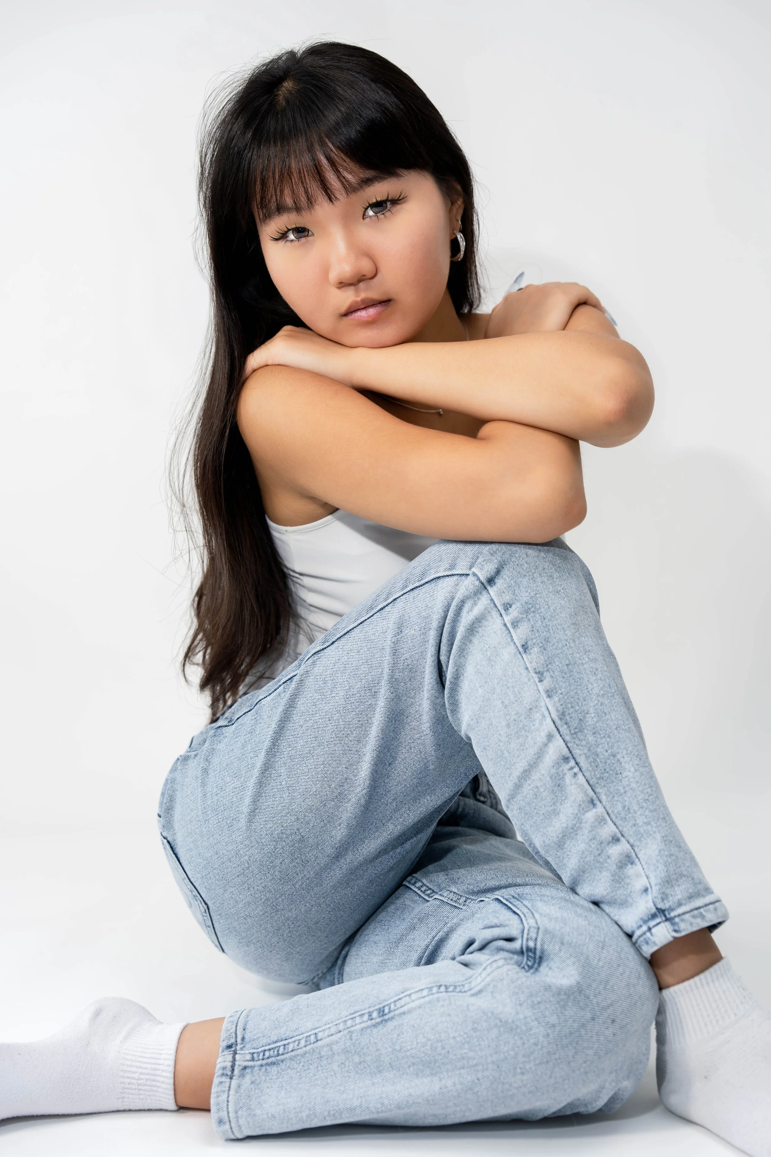 A young woman with long dark hair and bangs, wearing hoop earrings, a white tank top, and light blue jeans, sitting against a plain white background, looking at the camera with a neutral expression, with her arms resting on her knees.
