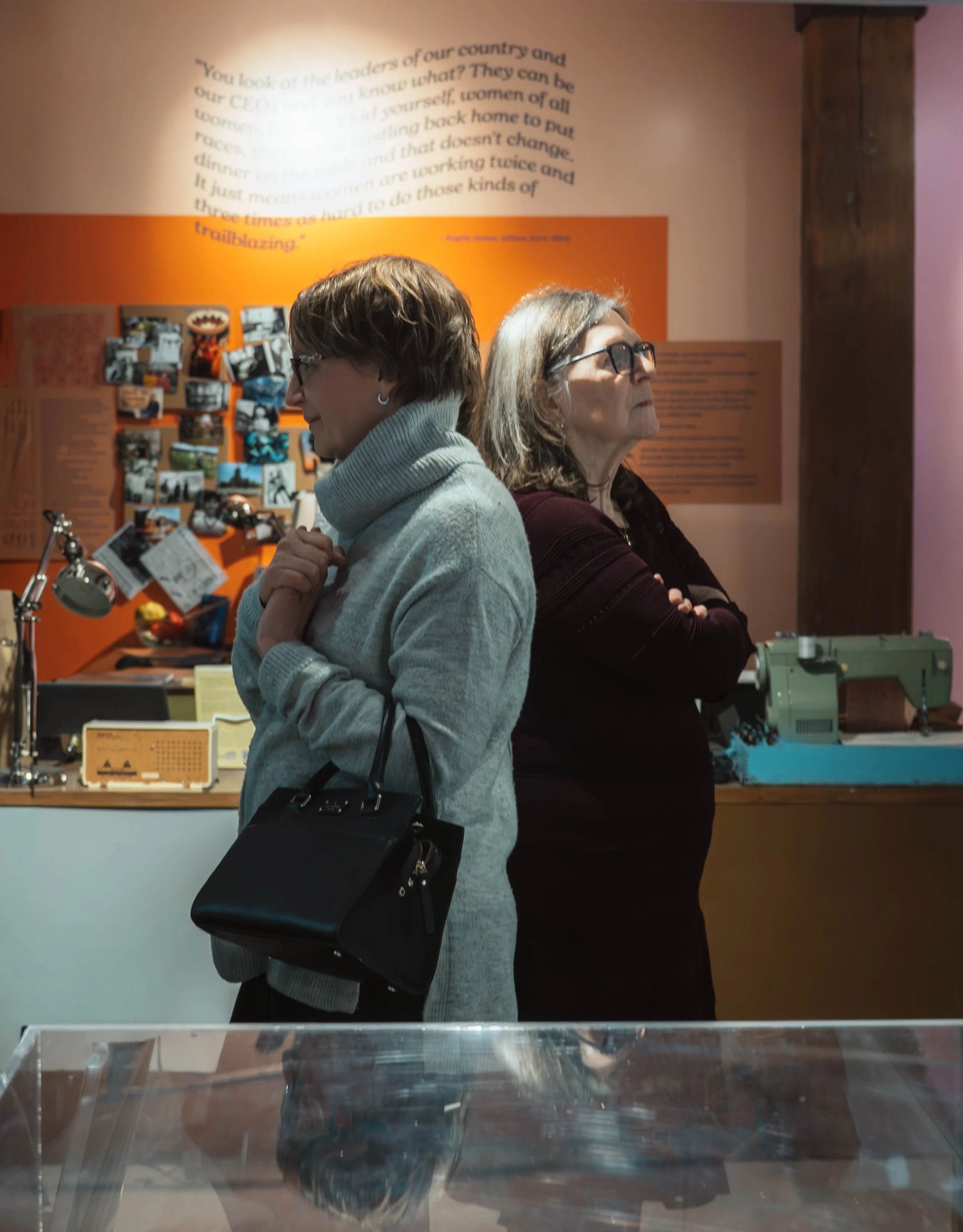 Two women standing back-to-back in a museum exhibit, with a display of photos and text on an orange background behind them.