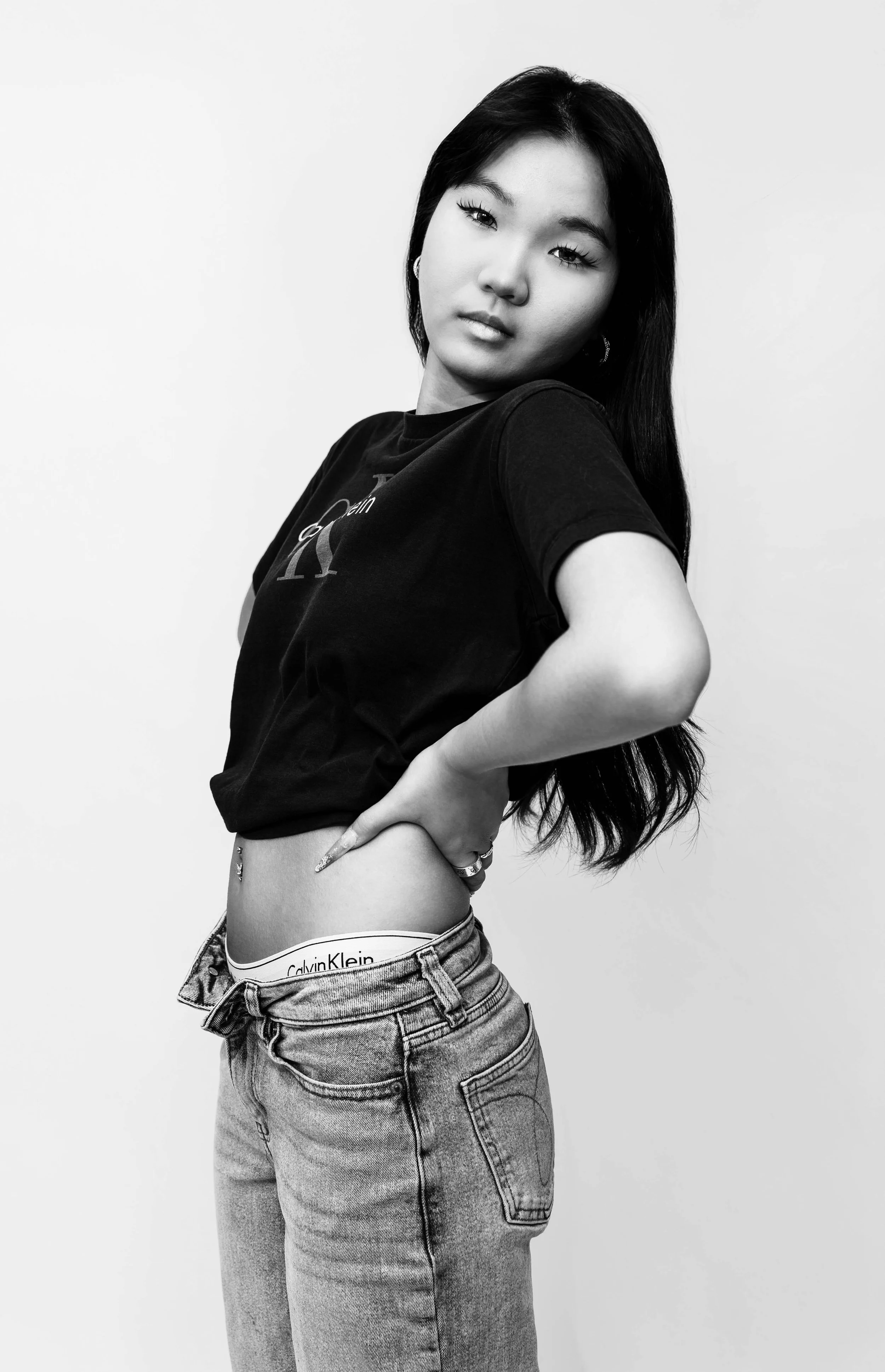 Black and white editorial studio portrait of Dana wearing a black Calvin Klein T-shirt and jeans, posing with one hand on her hip against a clean white backdrop, photographed by Lara Grace Creative Studio in Toronto.