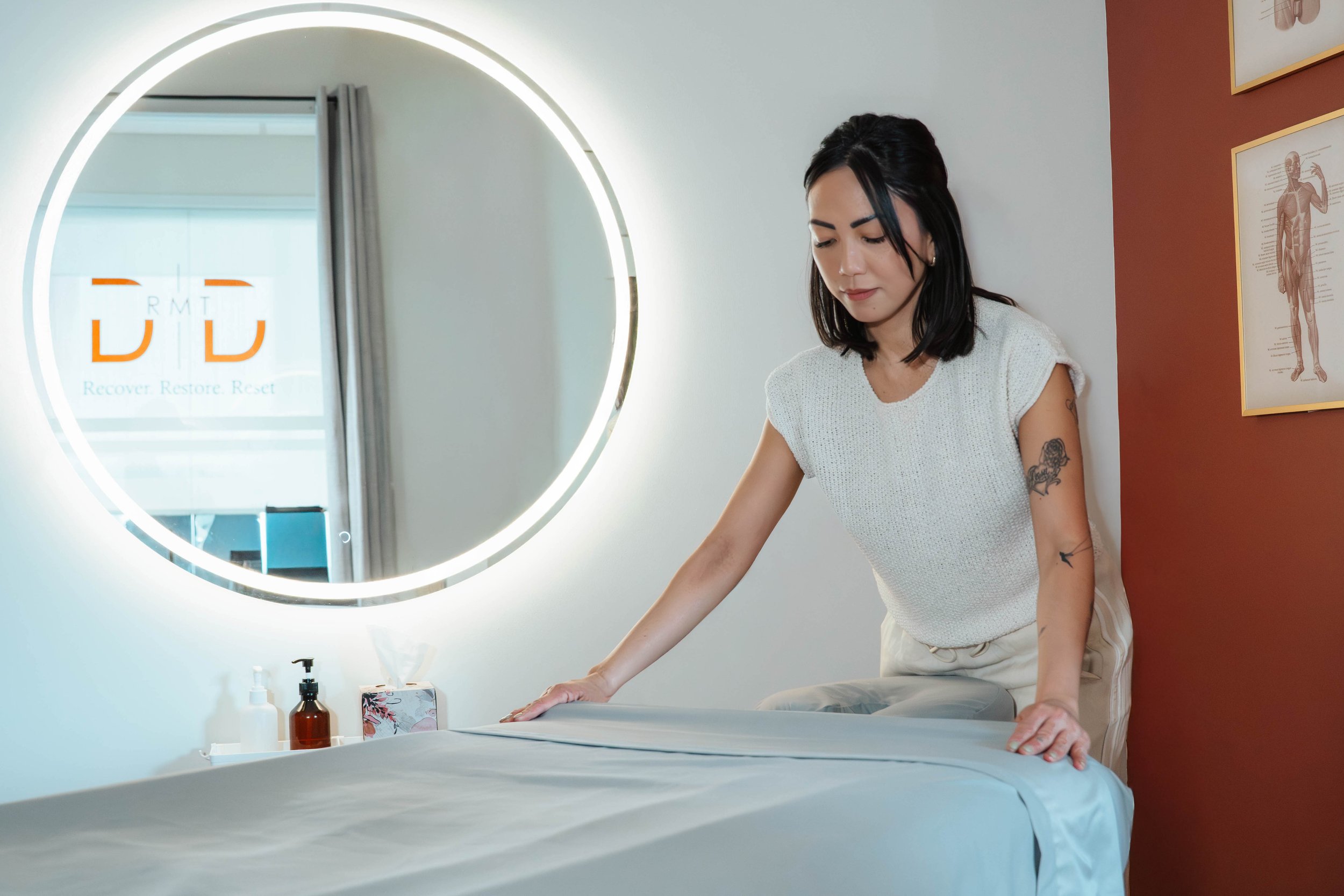 A woman with black hair doing massage therapy in a wellness center, standing at a massage table with her hands on it, near a round mirror with LED lighting, in a room with medical posters on the wall.