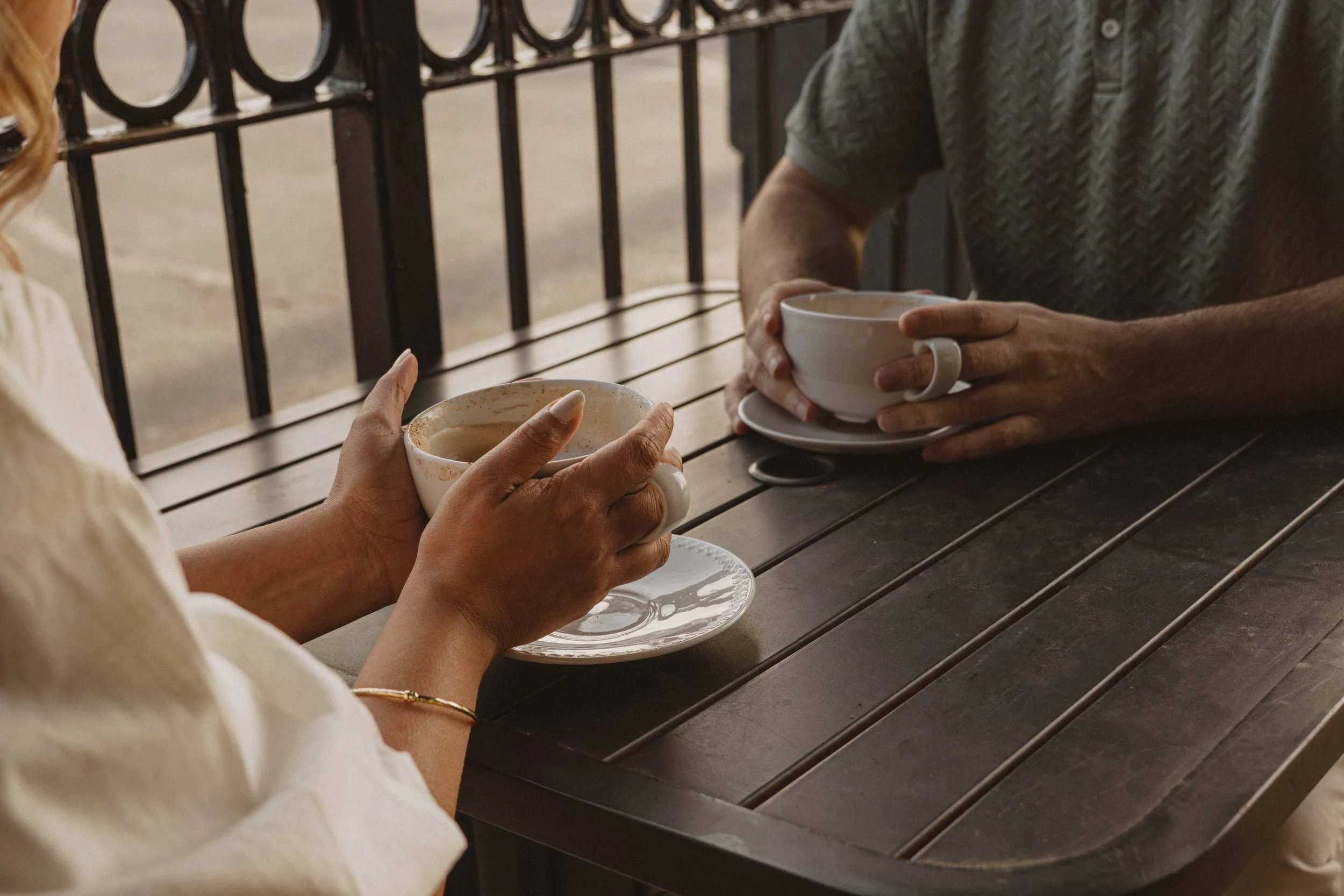 Two people are sitting at an outdoor table, each holding a cup of coffee or tea. One person's hand is visible with a ring, and they are wearing a white or cream-colored top. The other person is wearing a patterned shirt. The background includes a met