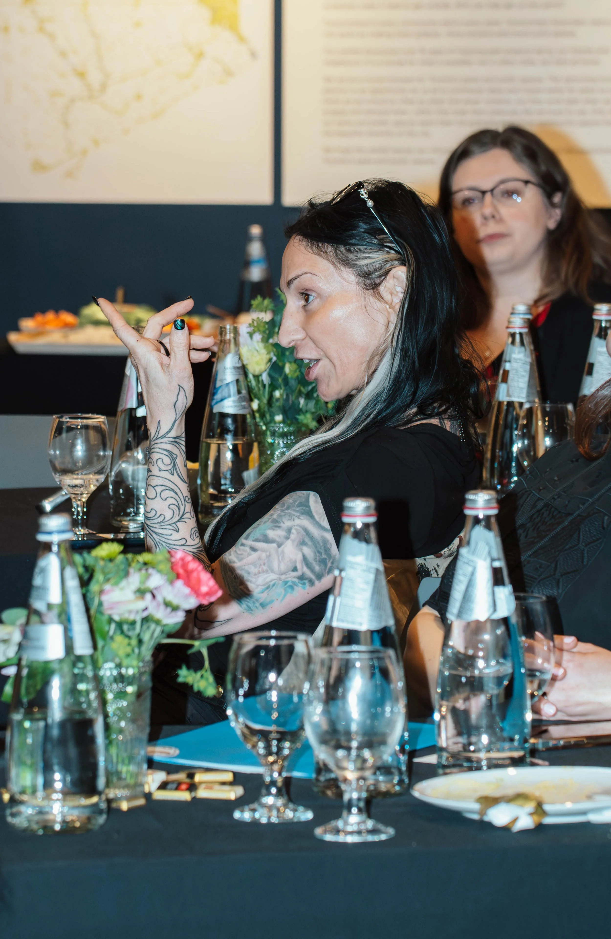 A woman with black and white hair, tattoos, and glasses on her head is sitting at a table during a dinner event, gesturing with her hand. There are bottles of water, glasses, flowers, and plates on the table. Another woman in glasses is sitting behin