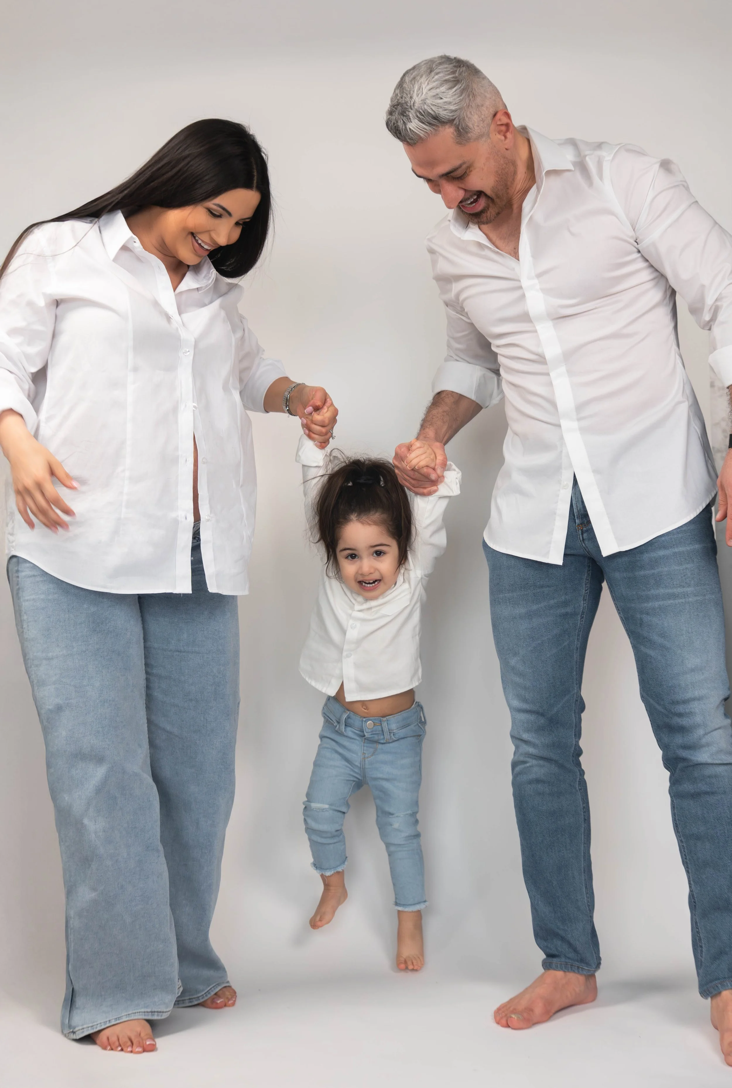A happy family with two parents and a girl holding hands and lifting her in the air against a plain white backdrop.