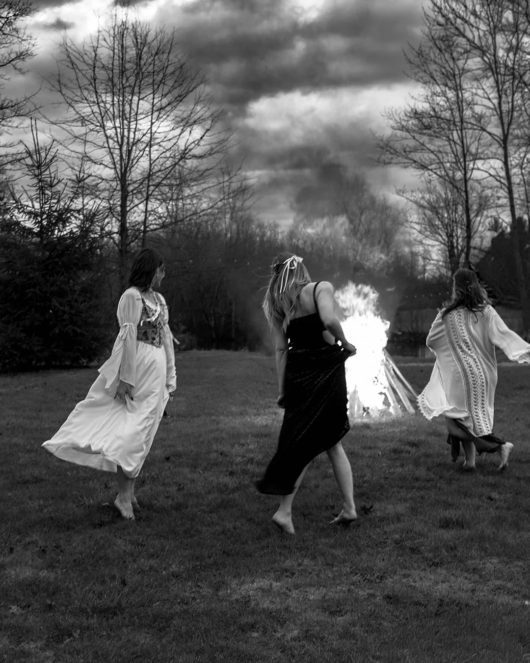 Three women in flowing dresses dancing around a bonfire on a grassy field during twilight, with bare trees and a cloudy sky in the background, in black and white.