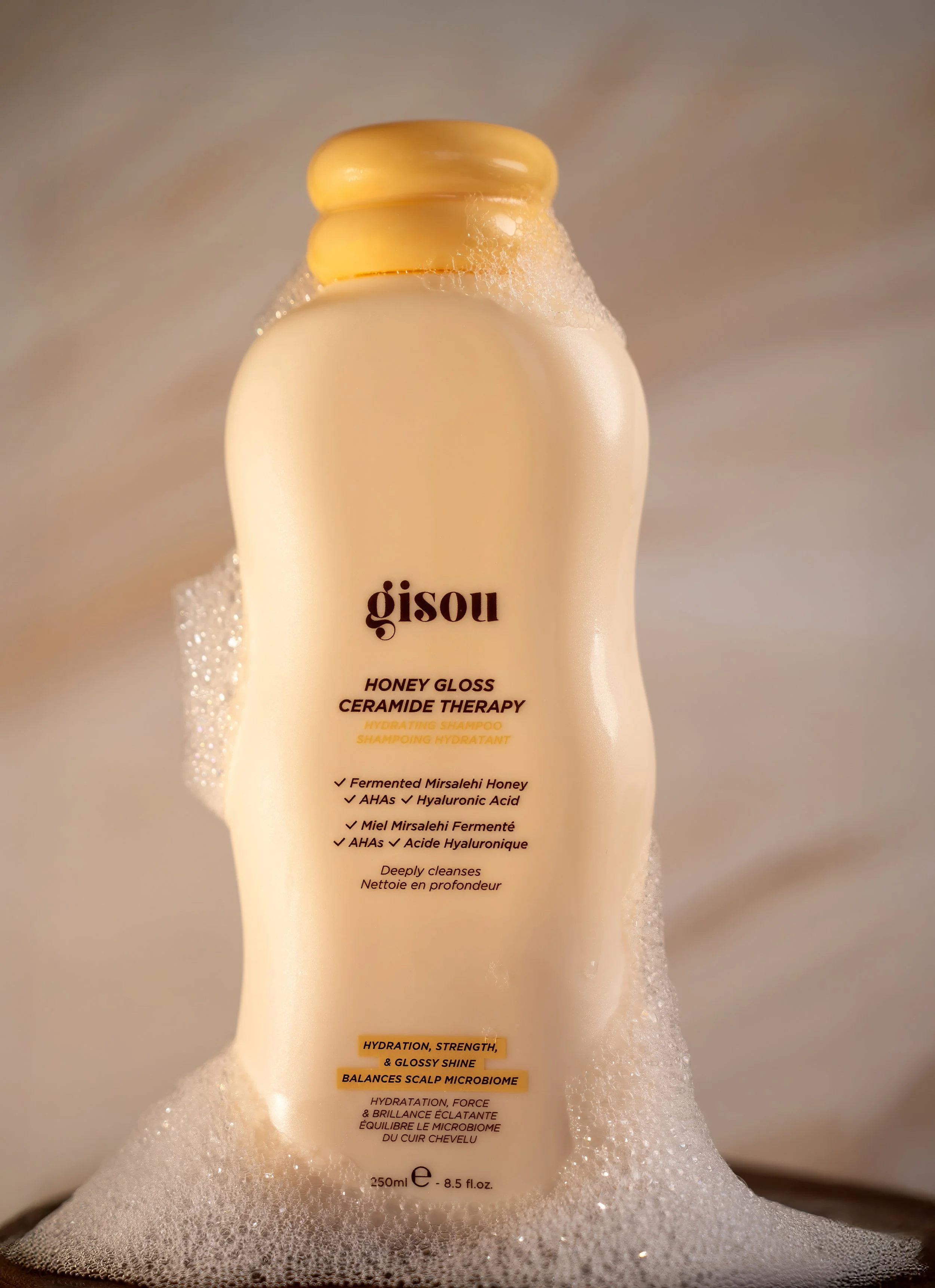Bottle of Gisou Honey Gloss Ceramide Therapy hydrating shampoo with foam and bubbles around it.