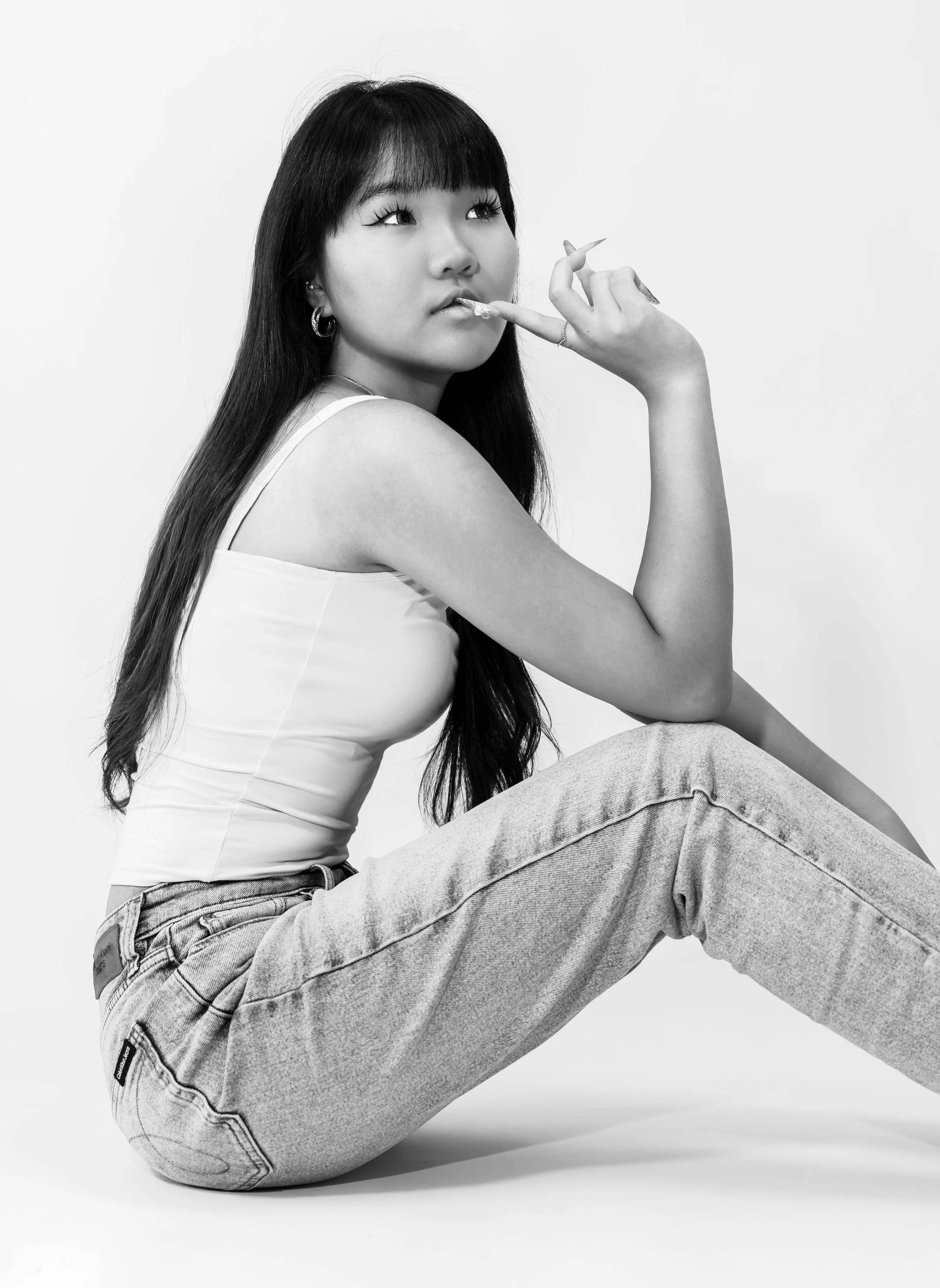 Black and white editorial studio portrait of Dana seated against a white backdrop, wearing a white tank top and jeans while posing with one hand near her lips and looking off camera, photographed by Lara Grace Creative Studio in Toronto.