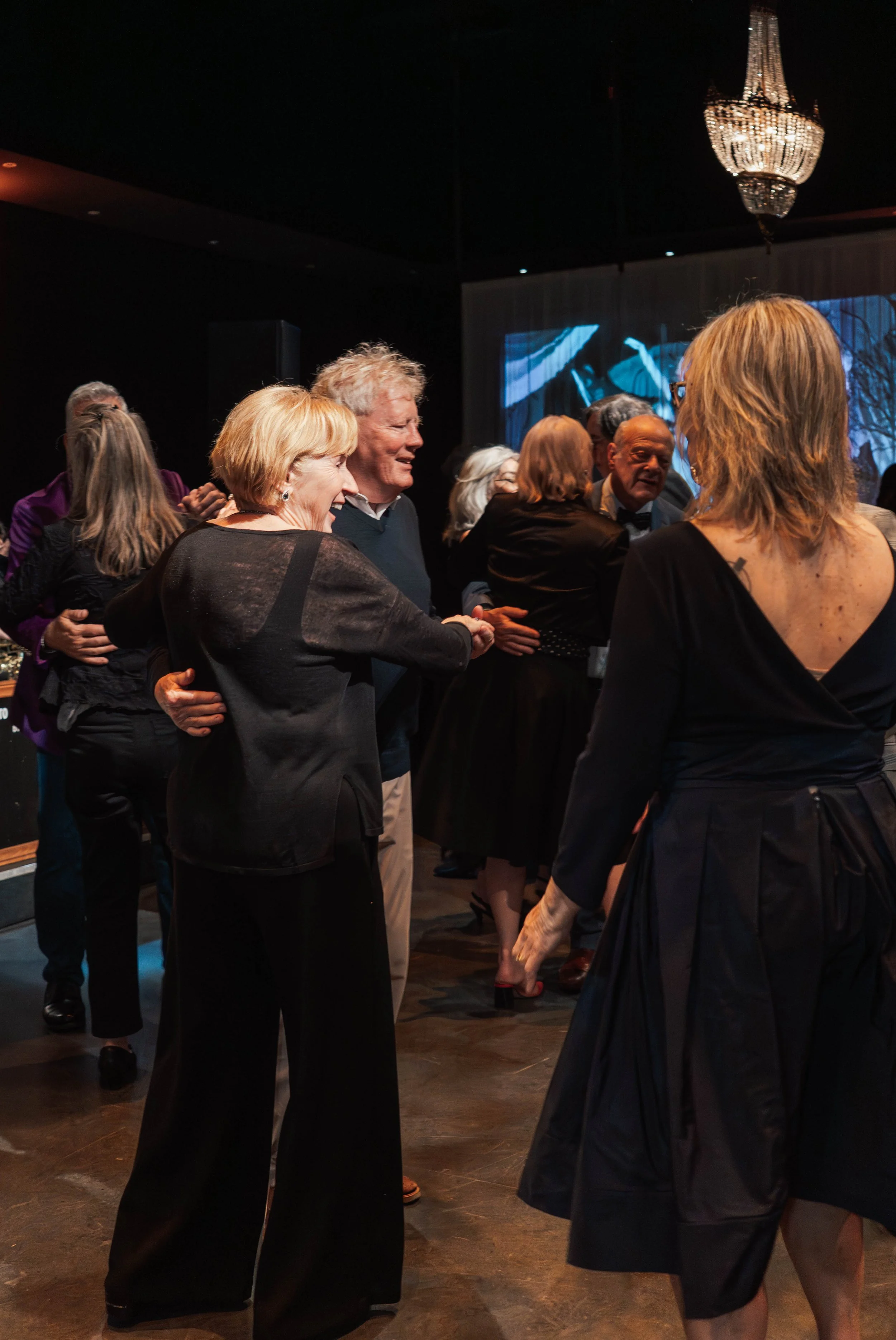 People dancing and socializing at a party or event inside a dimly lit room with a chandelier.