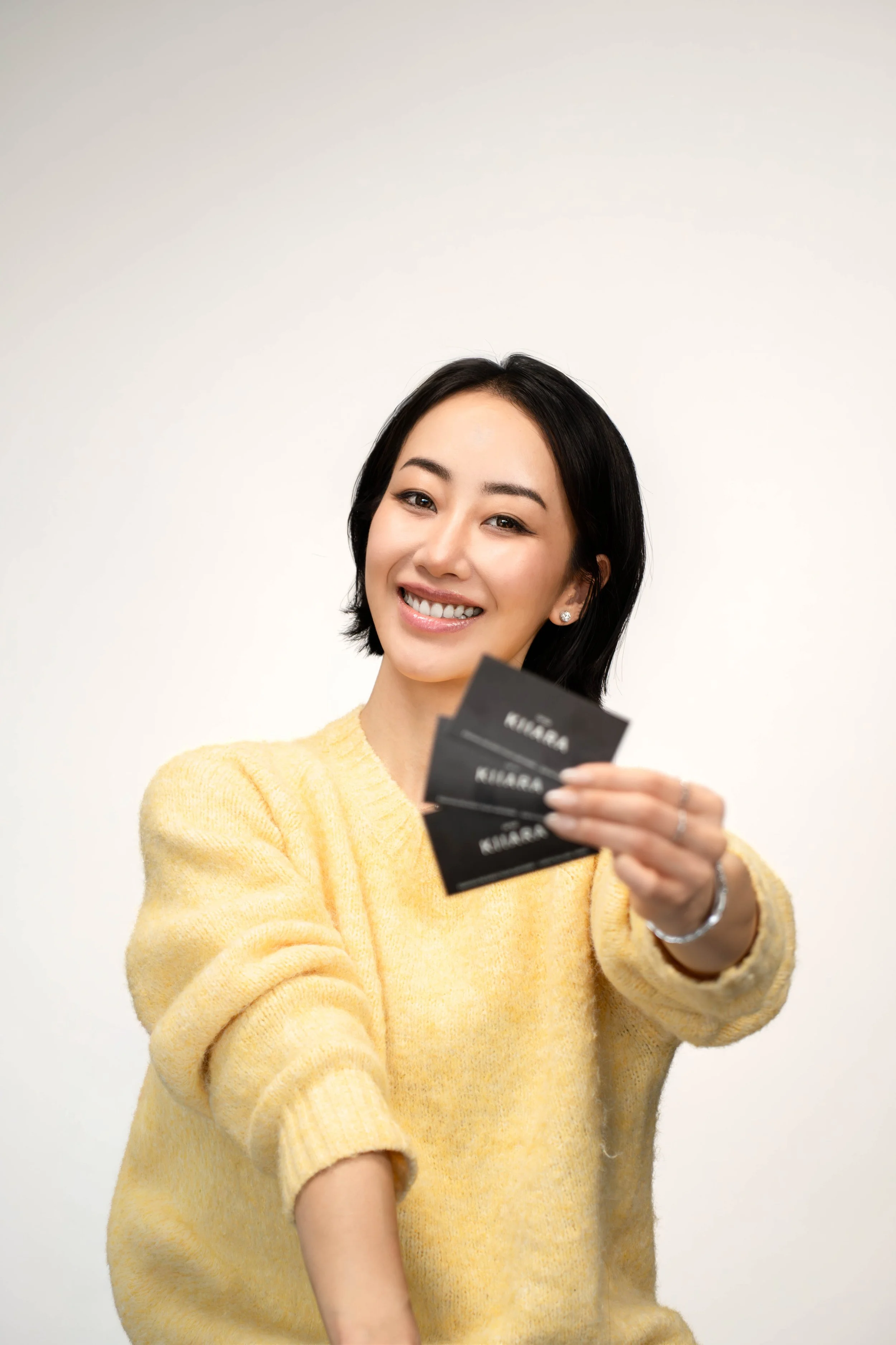 A woman with short black hair, wearing a yellow sweater, smiling and holding three black cards towards the camera.