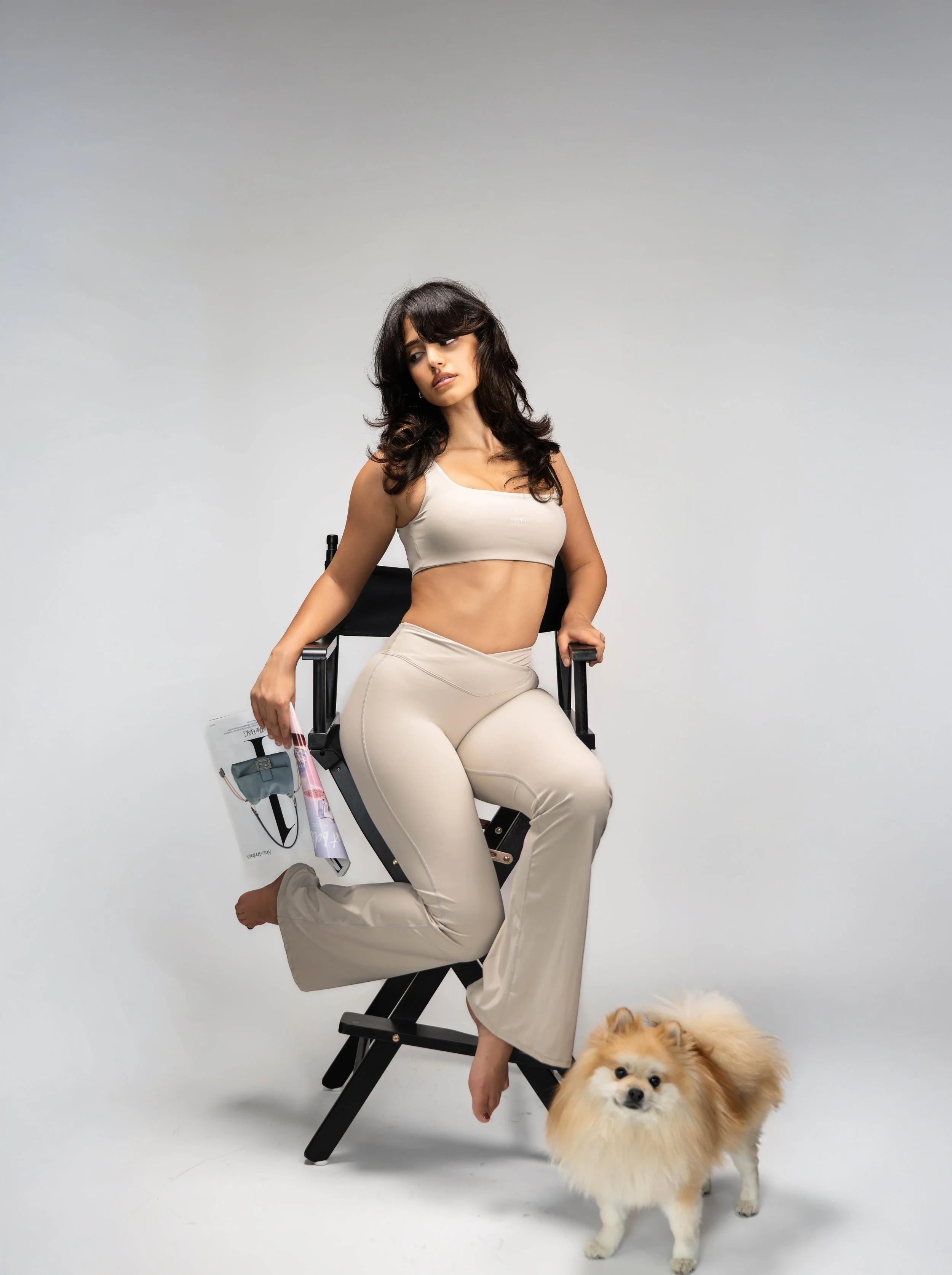 Woman sitting on a director's chair with a small fluffy dog standing next to her on a plain white background.
