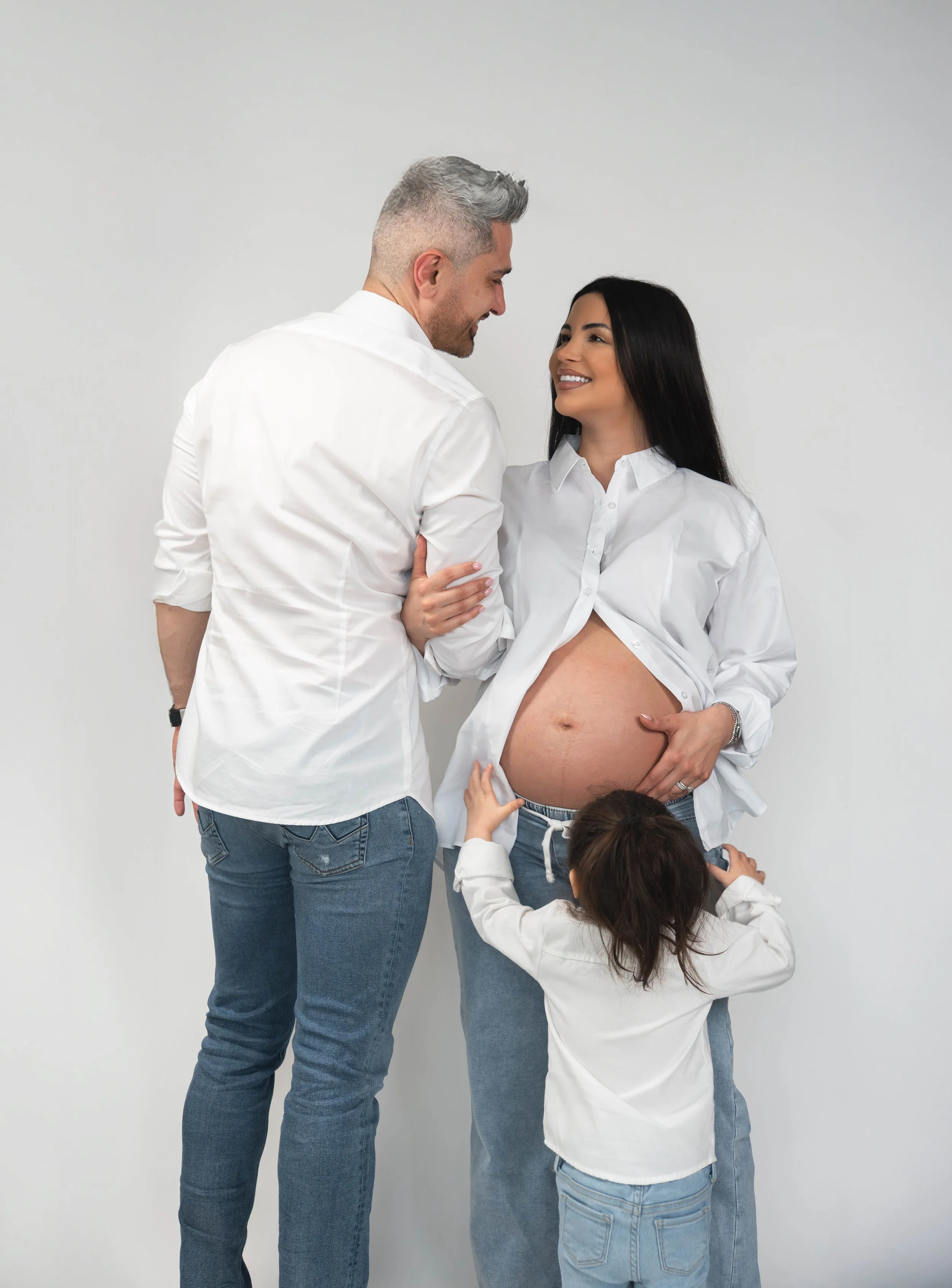 A pregnant woman with long dark hair wearing a white shirt and blue jeans, smiling and looking at a man. The man, with short gray hair and wearing a white shirt and blue jeans, is smiling and looking at the woman. There is also a young girl with brow