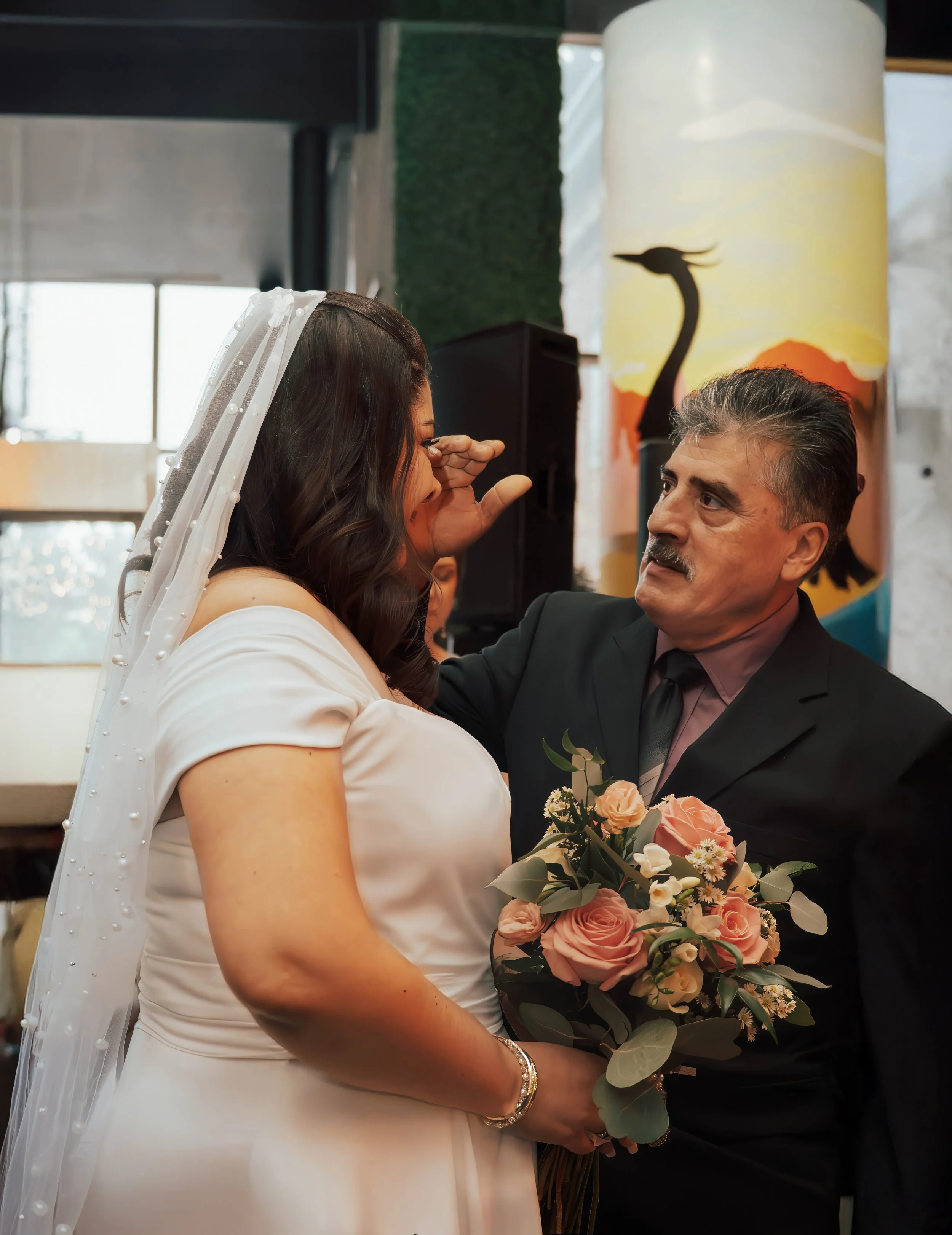 A bride holding a bouquet of pink roses and white flowers, standing face to face with an older man in a black suit, at a wedding reception.
