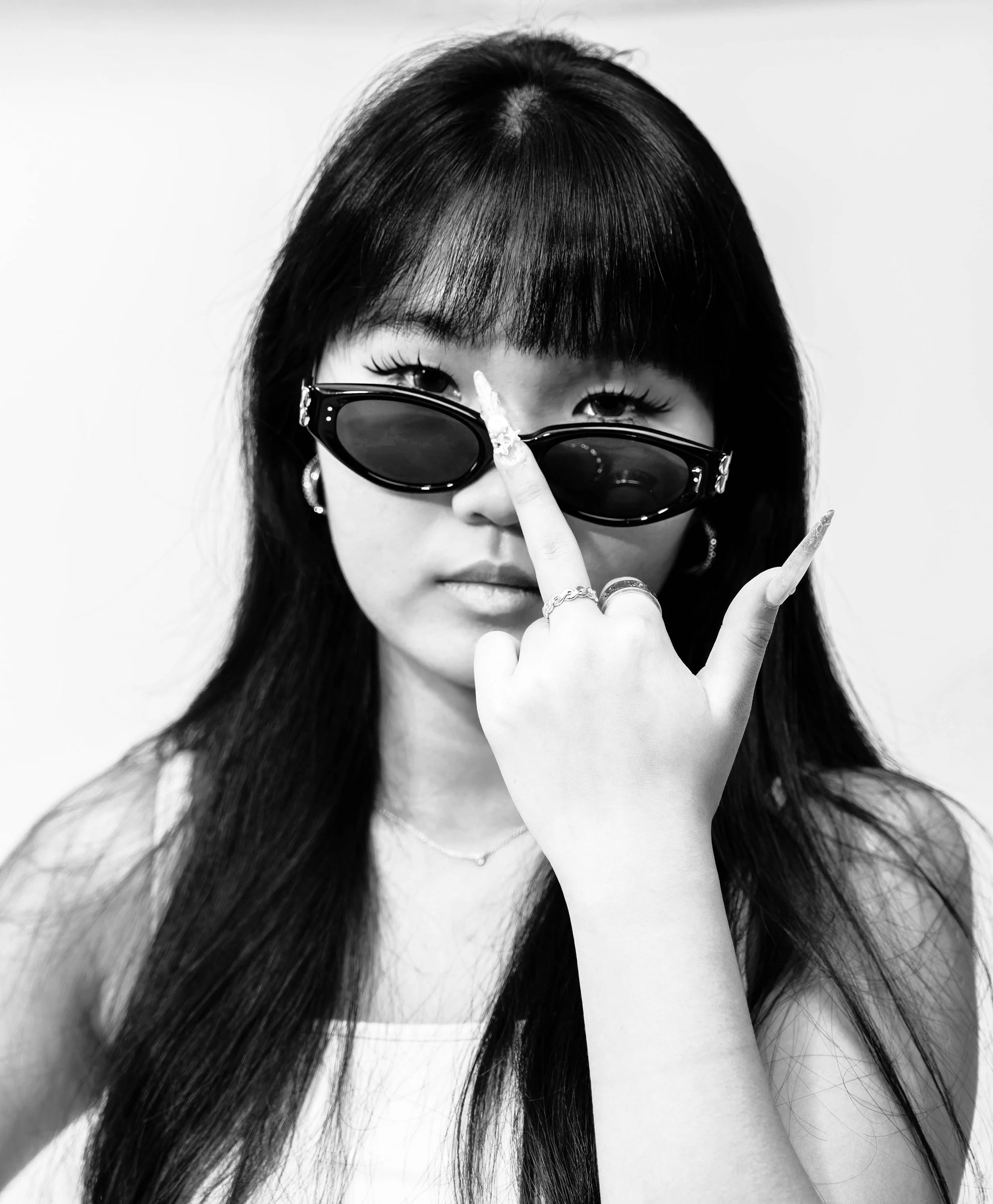 Black and white portrait of a woman with long dark hair, wearing sunglasses and jewelry, making a gesture with her hand near her face.
