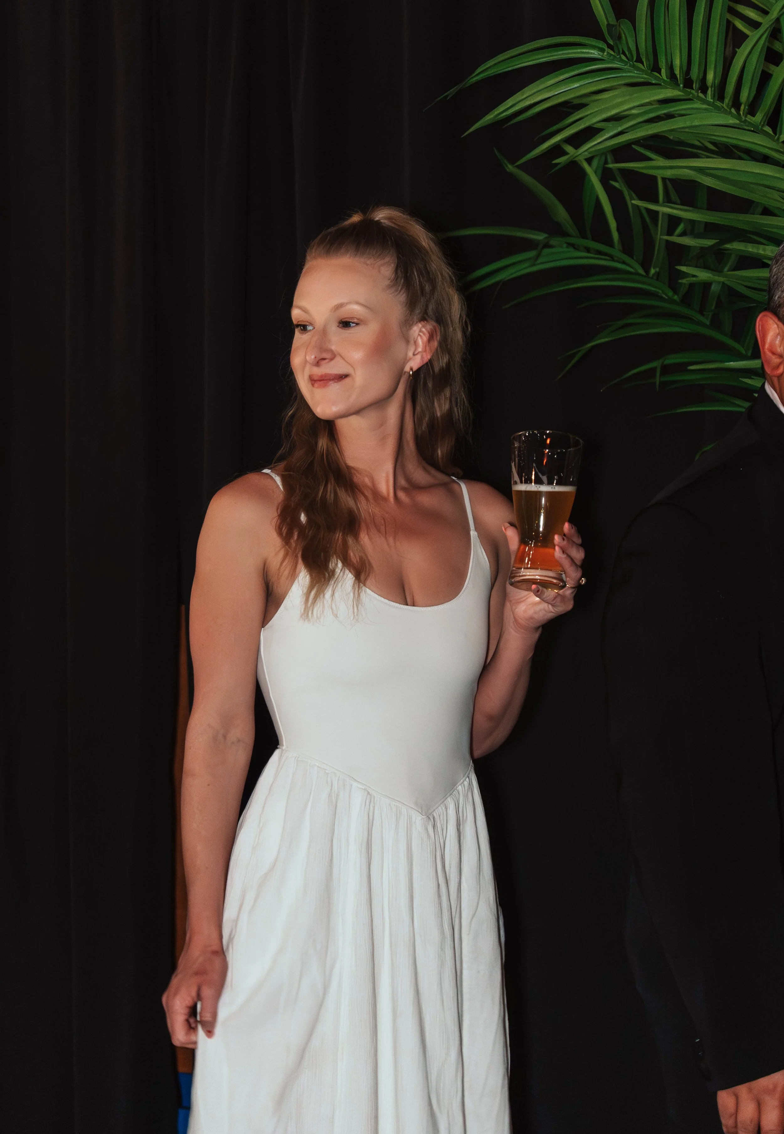 A woman in a white dress holding a glass of beer while standing beside a man in a black tuxedo, near green palm leaves in the background.