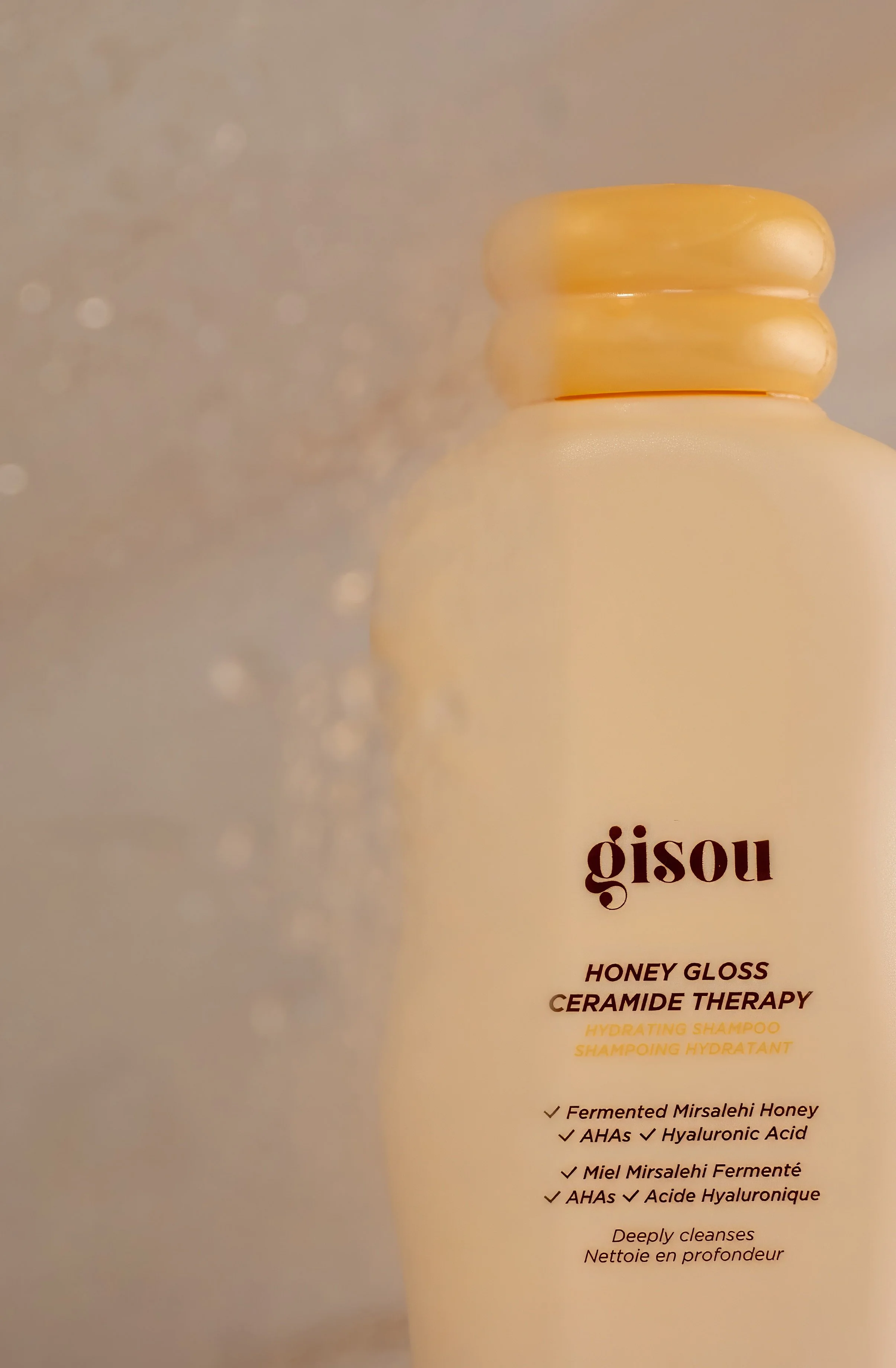 A beige bottle of Gisou Honey Gloss Ceramide Therapy Hydrating Shampoo with a gold cap. The label indicates it contains fermented Mirsalehi honey, AHAs, and hyaluronic acid, and states it deeply cleanses.