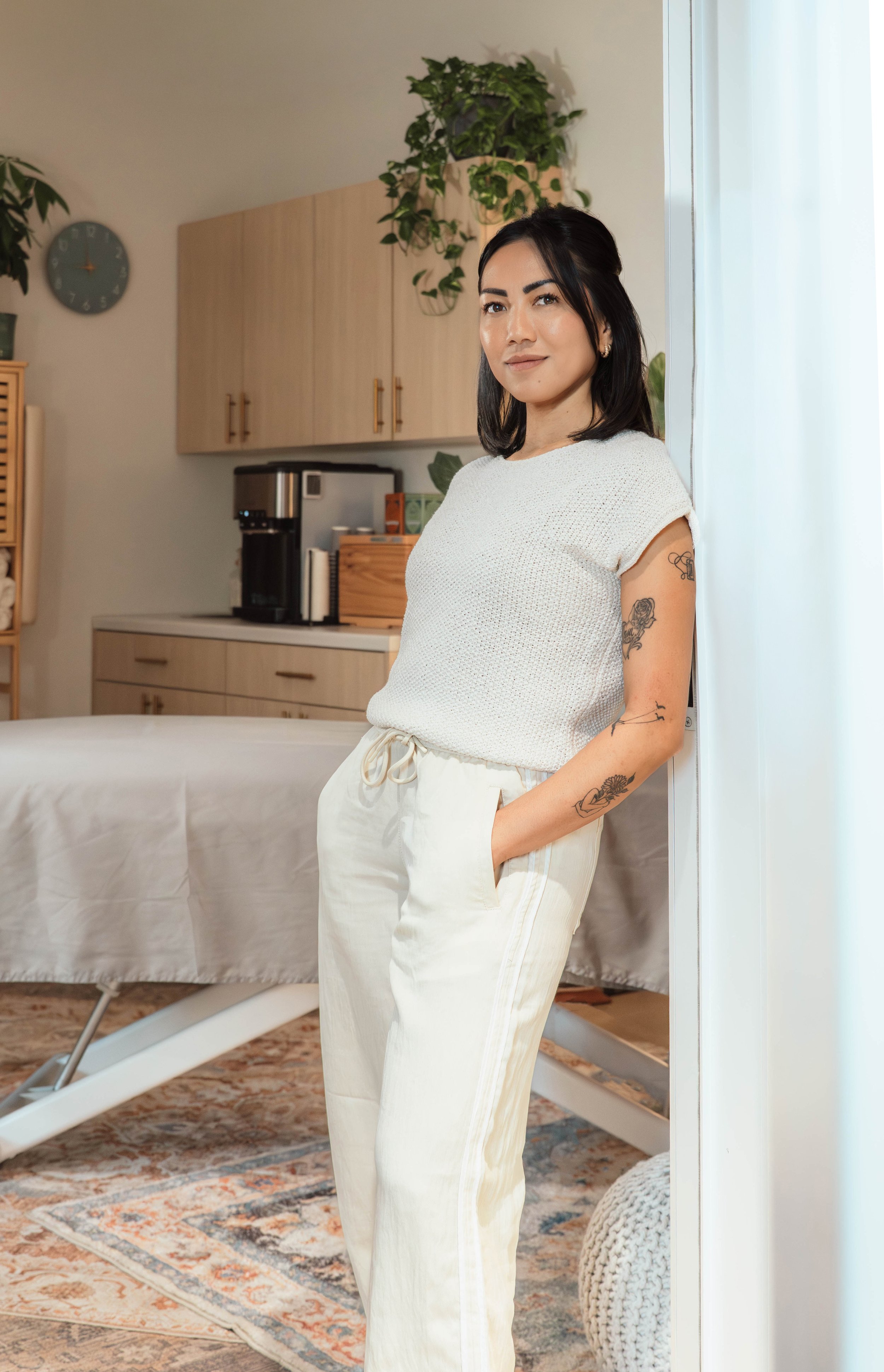 A woman with dark hair and tattoos on her arm, wearing a white top and white pants, standing in a room with a massage table, plants, a clock, and kitchen cabinets.